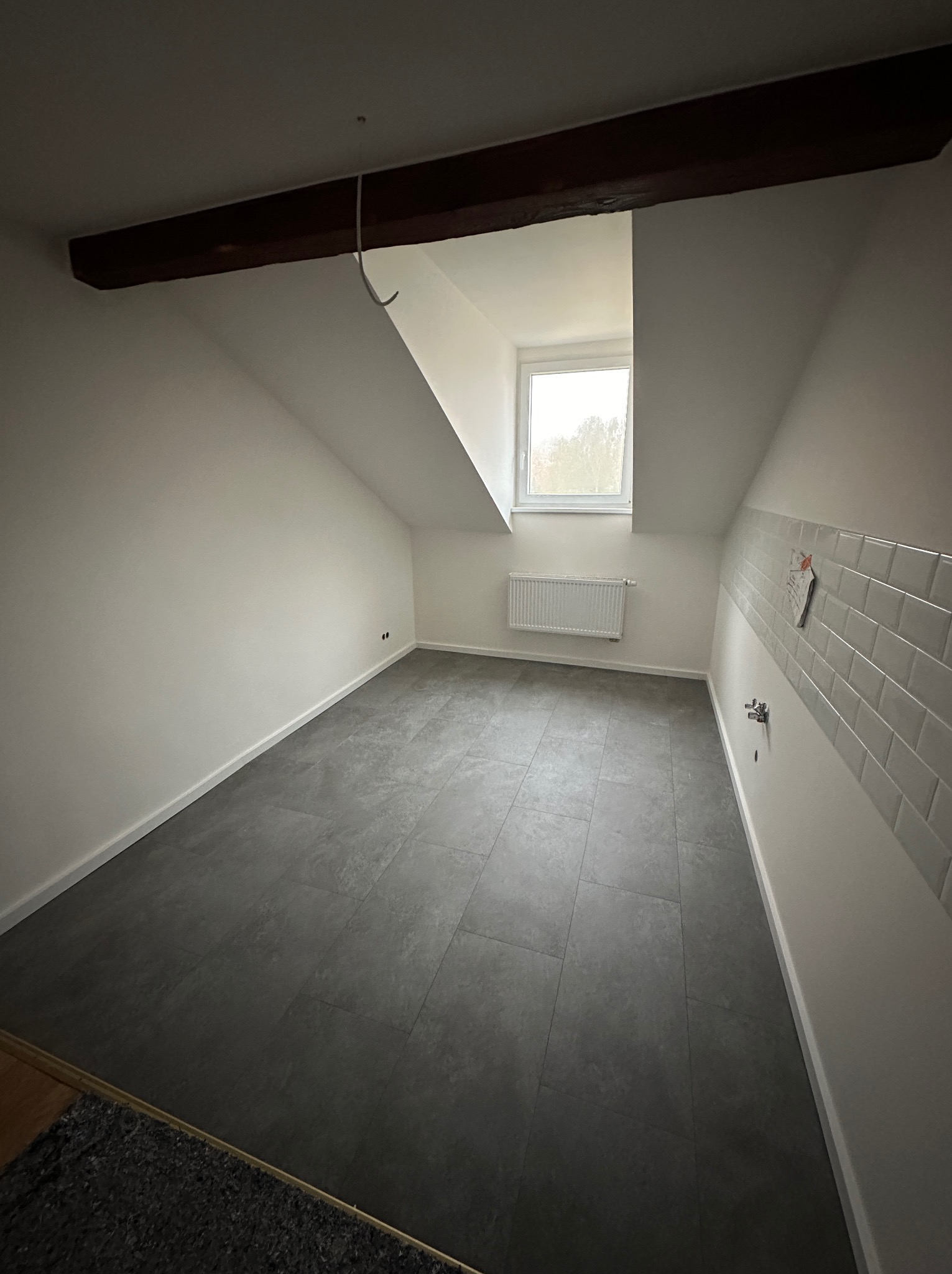 Poddasze po remoncie z szarą podłogą z płytek, świeżo pomalowanymi białymi ścianami, drewnianą belką stropową, białym grzejnikiem pod oknem i fragmentem ściany z białymi płytkami.