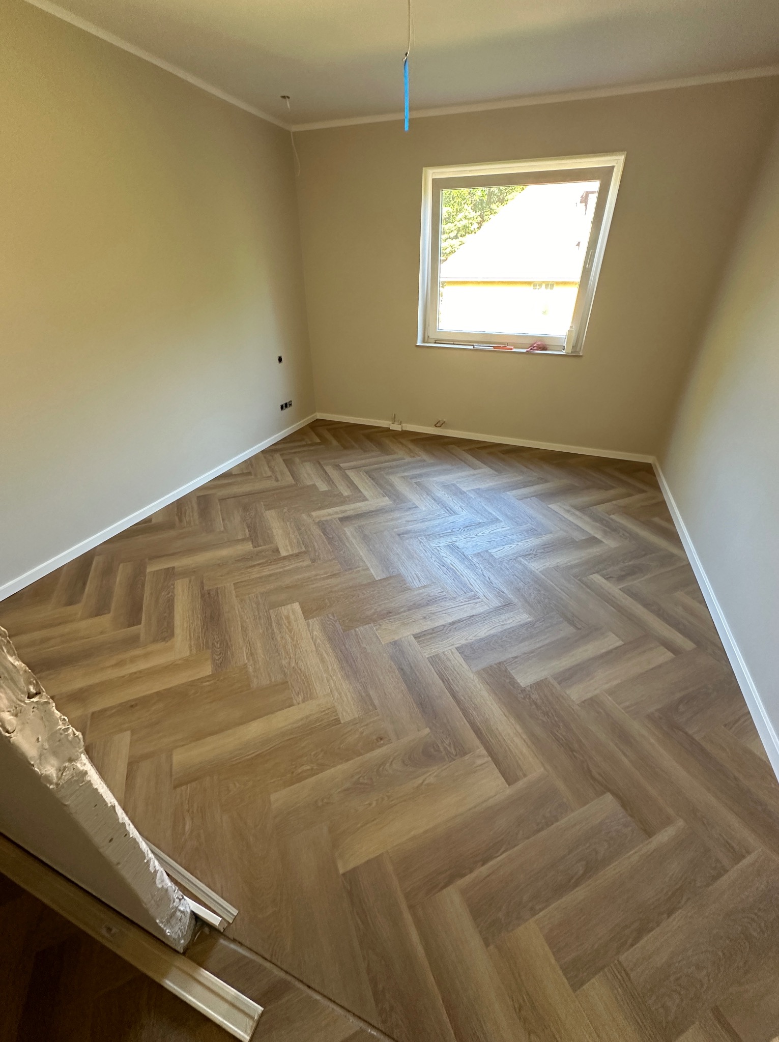 Pokój po remoncie z nowo ułożonymi panelami w jodełkę, widoczne listwy przypodłogowe i okno dachowe.