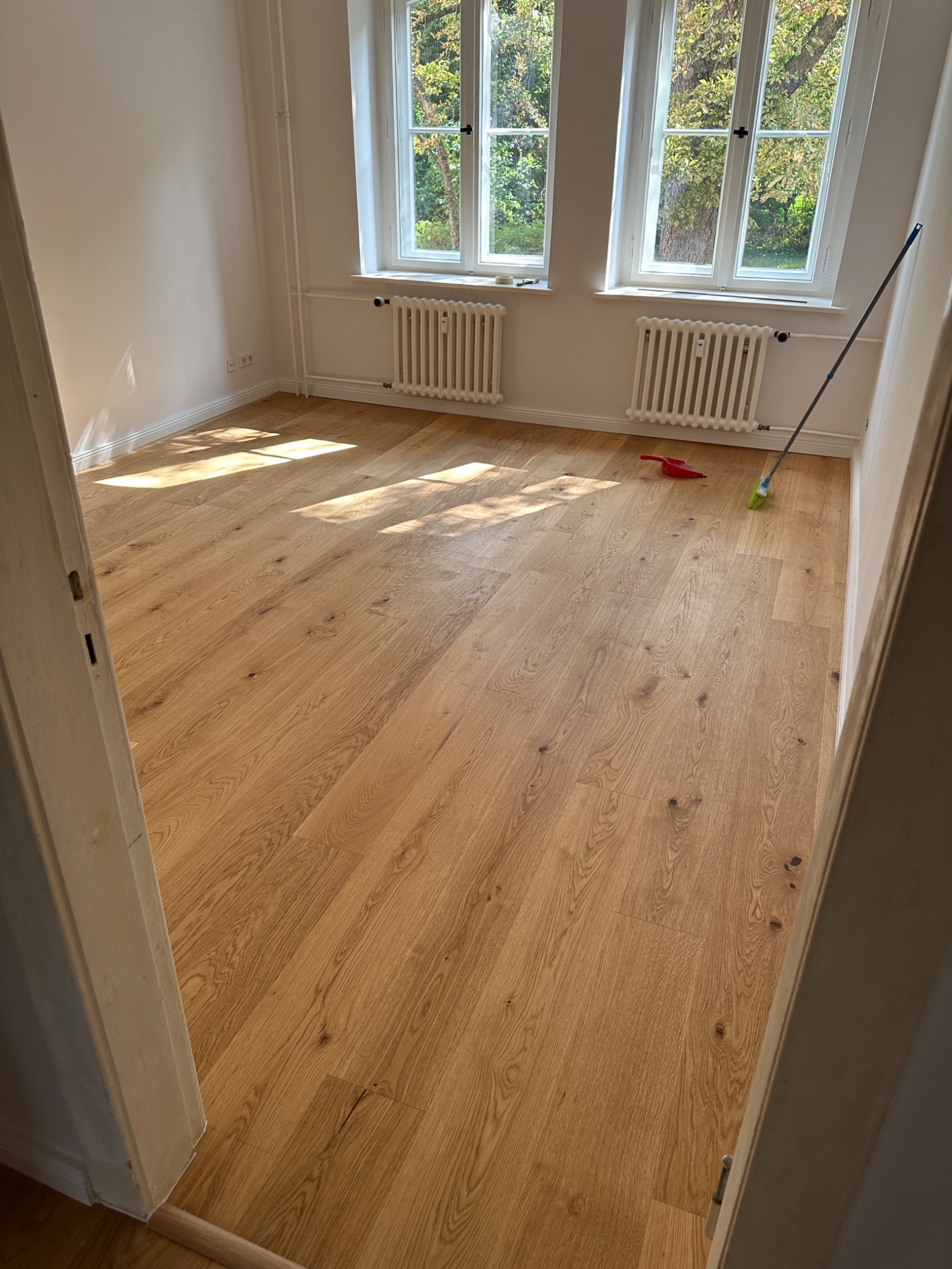 Jasne panele podłogowe ułożone w pokoju z dwoma oknami i białymi grzejnikami, widok zza otwartych drzwi, widoczna zmiotka i szufelka.