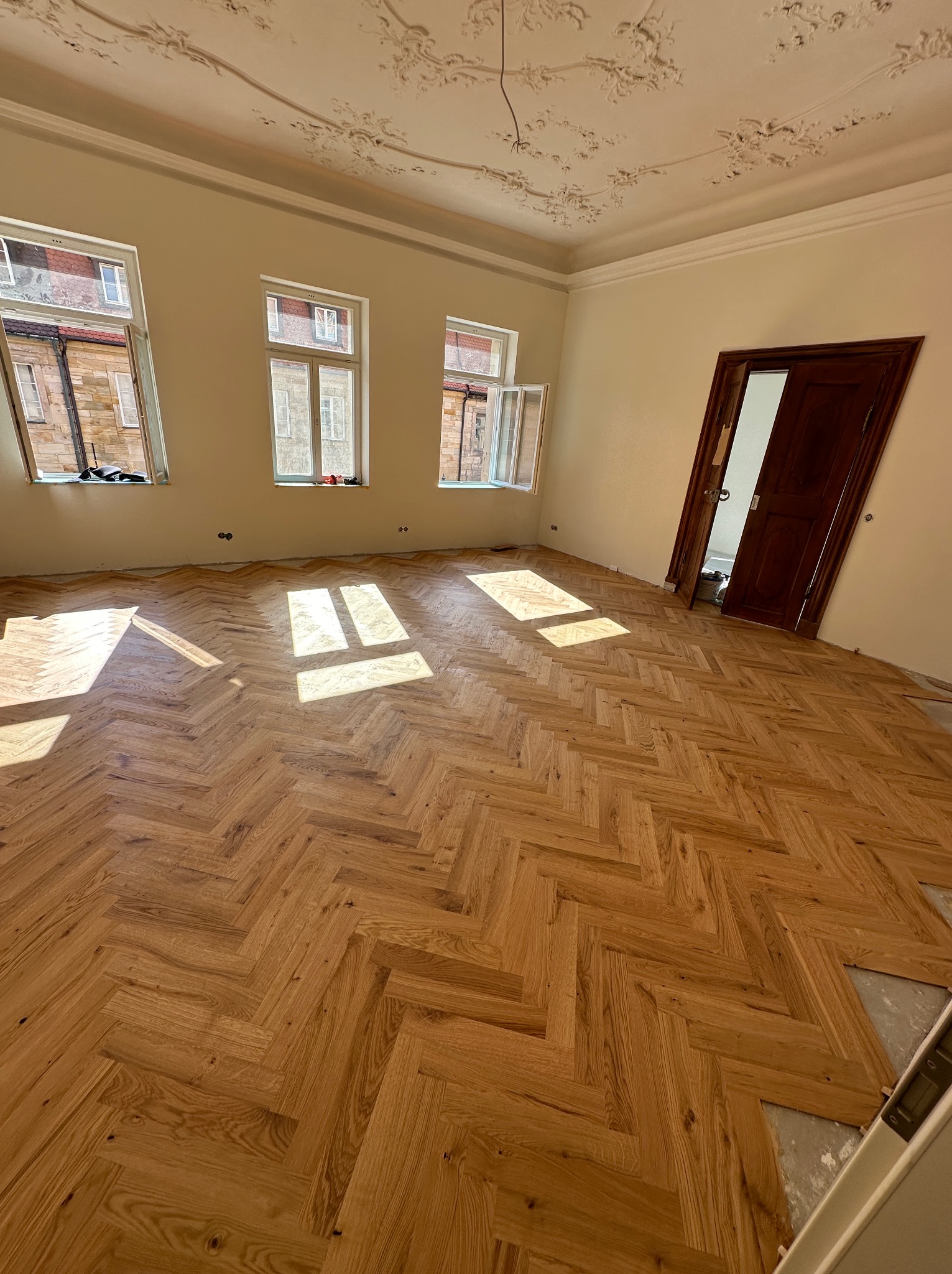 Nowo ułożony, jasny parkiet w jodełkę w przestronnym pokoju z trzema oknami i dekoracyjnym sufitem. Widoczne niedokończone fragmenty podłogi i fragment ściany przy drzwiach.