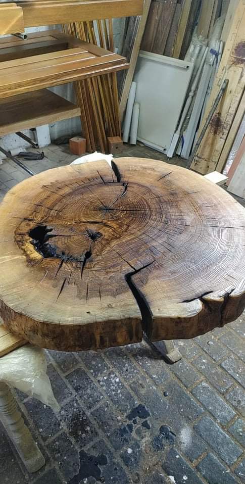 Stolik kawowy z plastra drewna z widocznymi pęknięciami i naturalnymi ubytkami, umieszczony w warsztacie z elementami drewnianymi w tle.