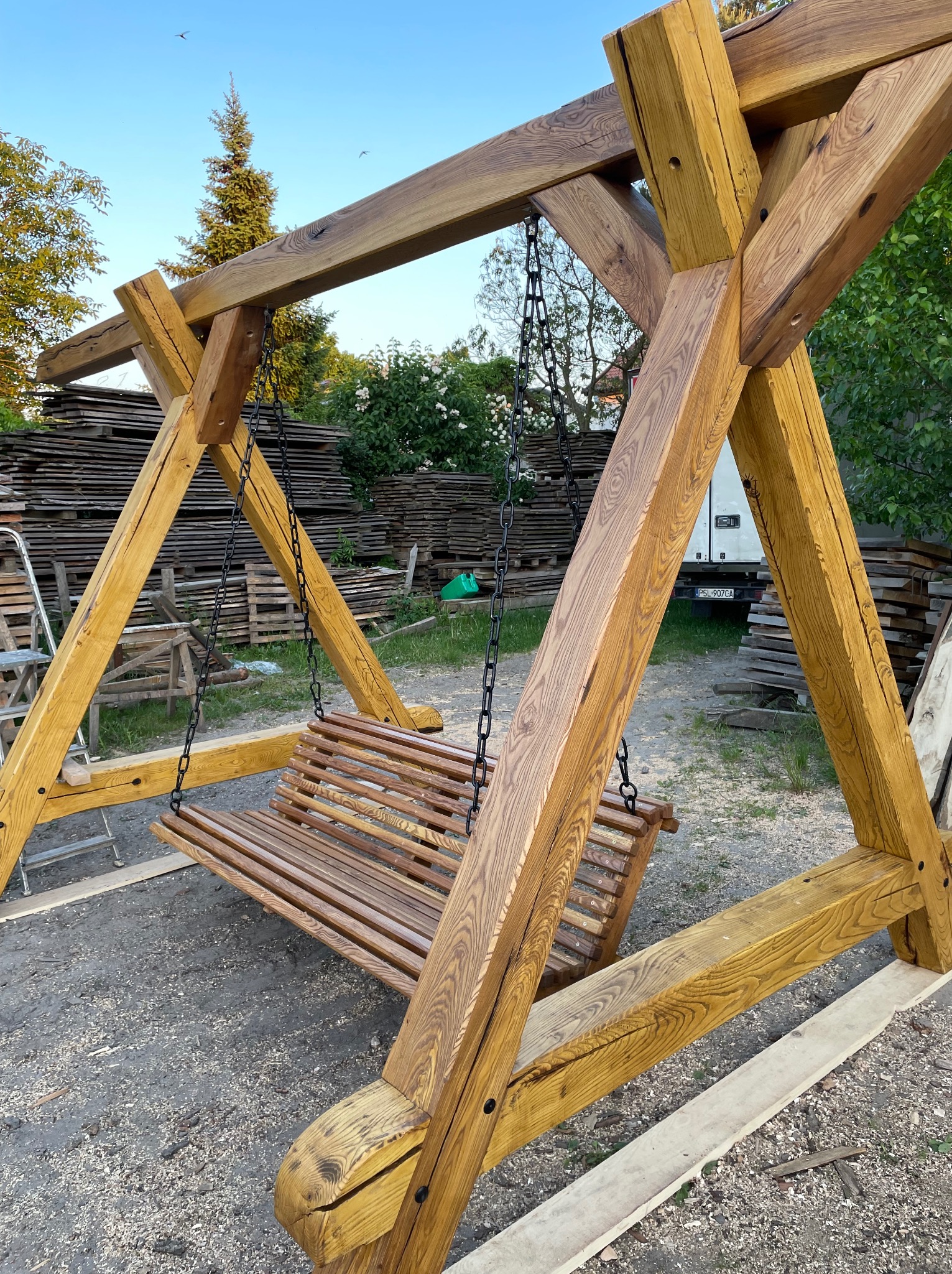 Drewniana huśtawka ogrodowa na łańcuchach z belek o wyraźnym usłojeniu, tło z ułożonych desek i białego busa.