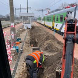 BUDLAT - Maciej Gajowniczek - Pracownik w pomarańczowej kamizelce kopie wąski rów na peronie kolejowym, obok stoi minikoparka, w tle widoczny pociąg i słup oświetleniowy.