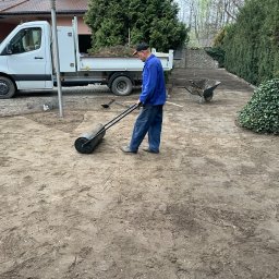 BUDLAT - Maciej Gajowniczek - Pracownik wyrównuje teren walcem ogrodowym przed ułożeniem trawy. W tle biała ciężarówka z ziemią i taczka. Wokół rosną tuje i krzewy.