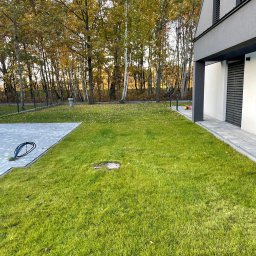 BUDLAT - Maciej Gajowniczek - Nowoczesny dom z zadbanym trawnikiem z rolki, widoczne przyłącze kablowe i jesienne drzewa w tle. Estetyczne zagospodarowanie przestrzeni.