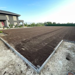 BUDLAT - Maciej Gajowniczek - Przygotowany teren pod trawnik z obrzeżami, widoczny pracownik i budynek w tle. Świeżo nawieziona ziemia, gotowa do położenia trawy z rolki. Ujęcie z bliska.