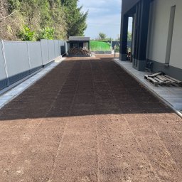 BUDLAT - Maciej Gajowniczek - Przygotowany teren pod trawnik z widocznymi śladami po bronowaniu, obrzeża z kostki, szara palisada i budynek z elementami grafitowymi w tle.