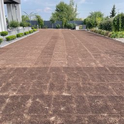 BUDLAT - Maciej Gajowniczek - Przygotowany teren pod trawnik z widocznymi śladami po wałowaniu, obrzeża z kostki i roślin, nowoczesny dom w tle, błękitne niebo z chmurami.