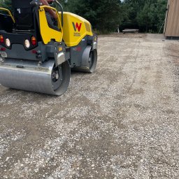 BUDLAT - Maciej Gajowniczek - Żółty walec Wacker Neuson RD28 ubija kamienistą nawierzchnię drogi w pobliżu lasu i budynku. Operator w pomarańczowej koszulce siedzi na maszynie.