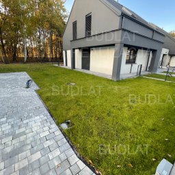 BUDLAT - Maciej Gajowniczek - Nowoczesny dom z szarą elewacją i zadbanym trawnikiem. Widoczny fragment brukowanej ścieżki i jesienne drzewa w tle. Ujęcie z poziomu gruntu.