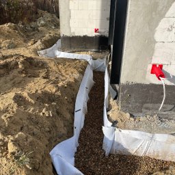 BUDLAT - Maciej Gajowniczek - Wykop fundamentowy zabezpieczony geowłókniną i wypełniony żwirem, widoczne fragmenty ścian budynku w trakcie budowy, instalacja elektryczna na zewnątrz.