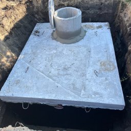 BUDLAT - Maciej Gajowniczek - Betonowa płyta szamba w wykopie, z otwartym włazem. Widoczne haki do podnoszenia płyty i świeże uszczelnienie włazu. Grunt wokół wykopu.