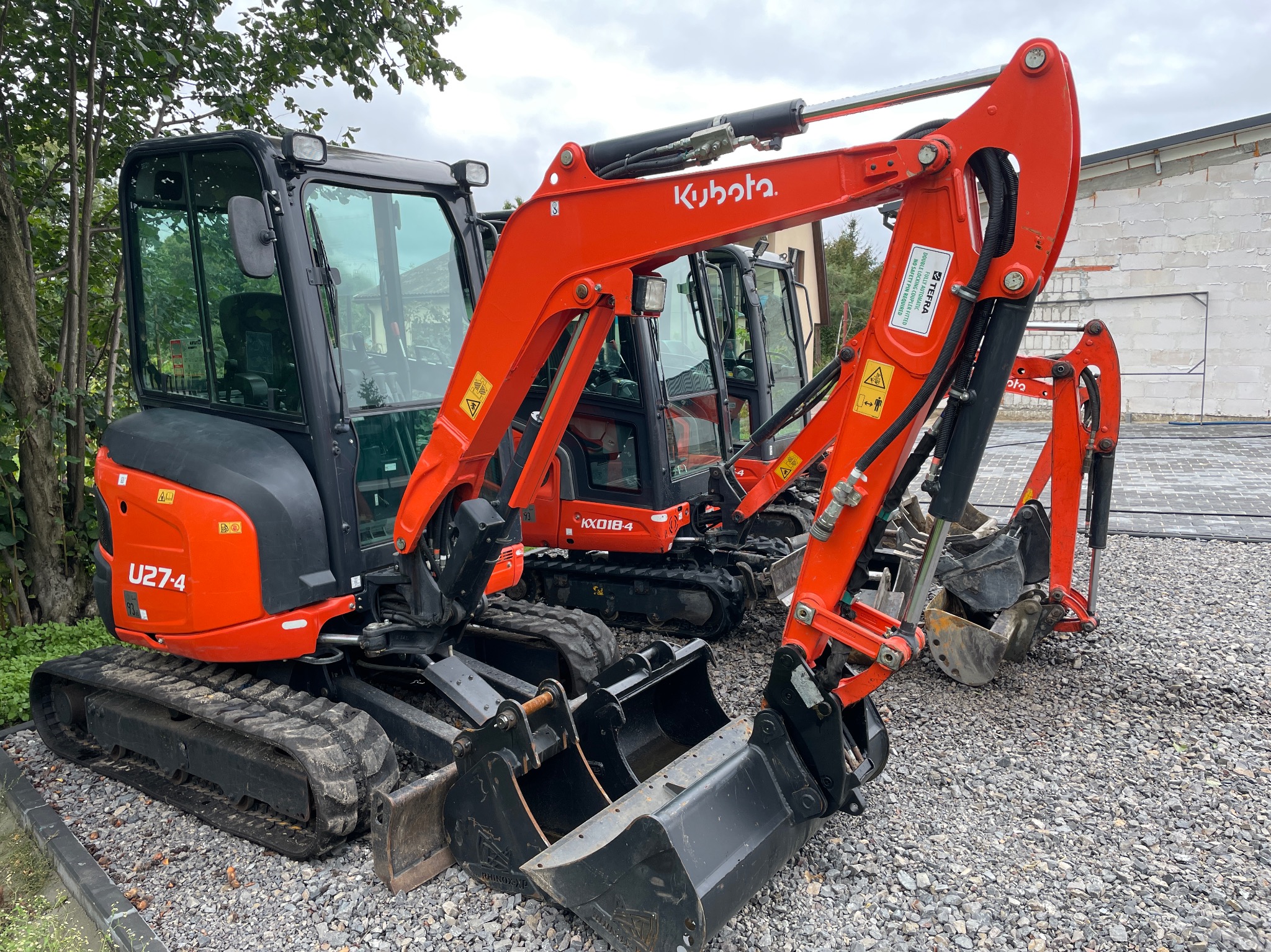 Pomarańczowe minikoparki Kubota U27-4 i KX018-4, z łyżkami, stoją na żwirowym terenie w Latowiczu, gotowe do robót ziemnych.