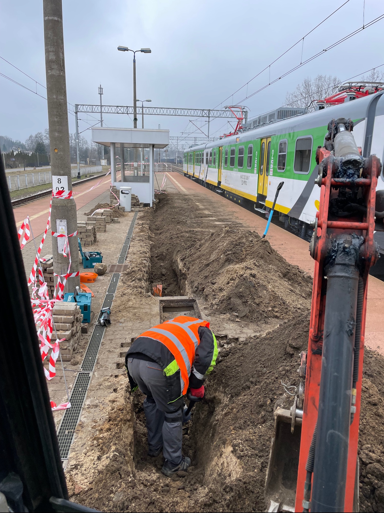 Pracownik w pomarańczowej kamizelce kopie wąski rów na peronie kolejowym, obok stoi minikoparka, w tle widoczny pociąg i słup oświetleniowy.