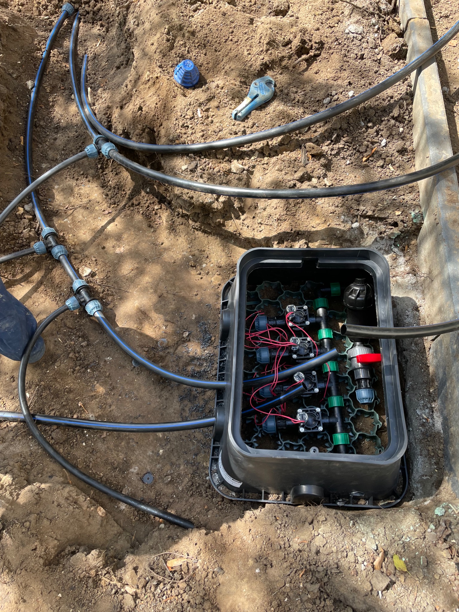 Instalacja systemu nawadniania w ziemi: czarne rury, złączki, zawory w plastikowej skrzynce. Widoczne narzędzie do cięcia rur i niebieska złączka.