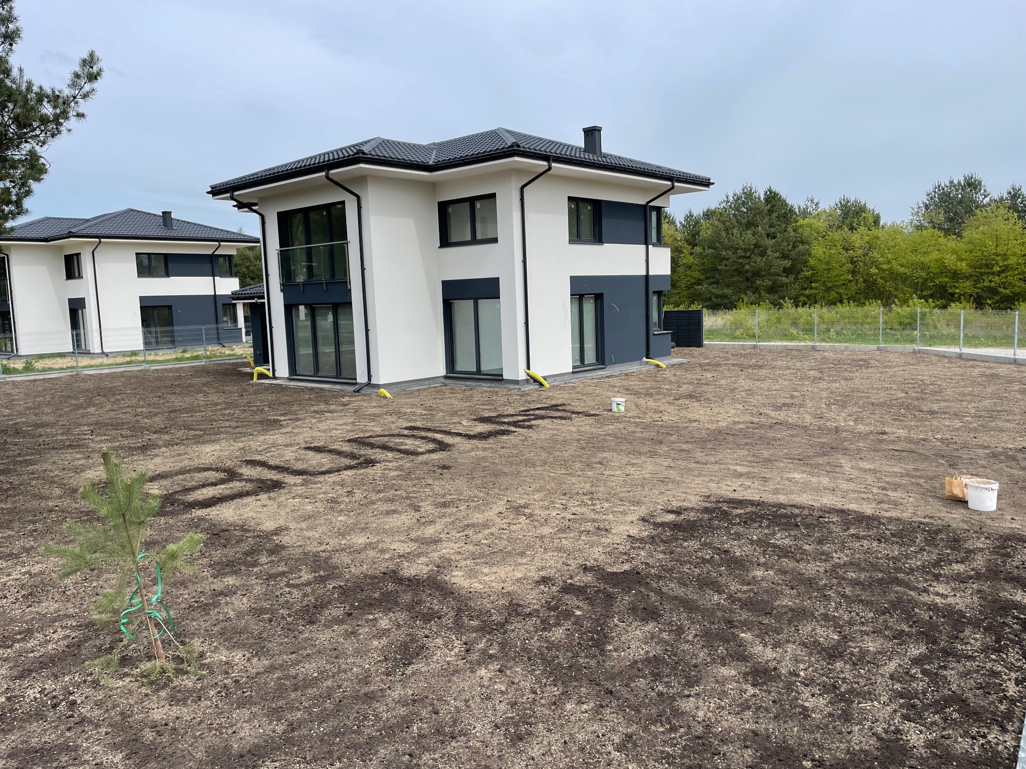 Nowoczesny dom z ogrodem w trakcie zakładania, widoczne napisy w ziemi i małe drzewko. Szaro-biała elewacja, ogrodzenie z siatki w tle.