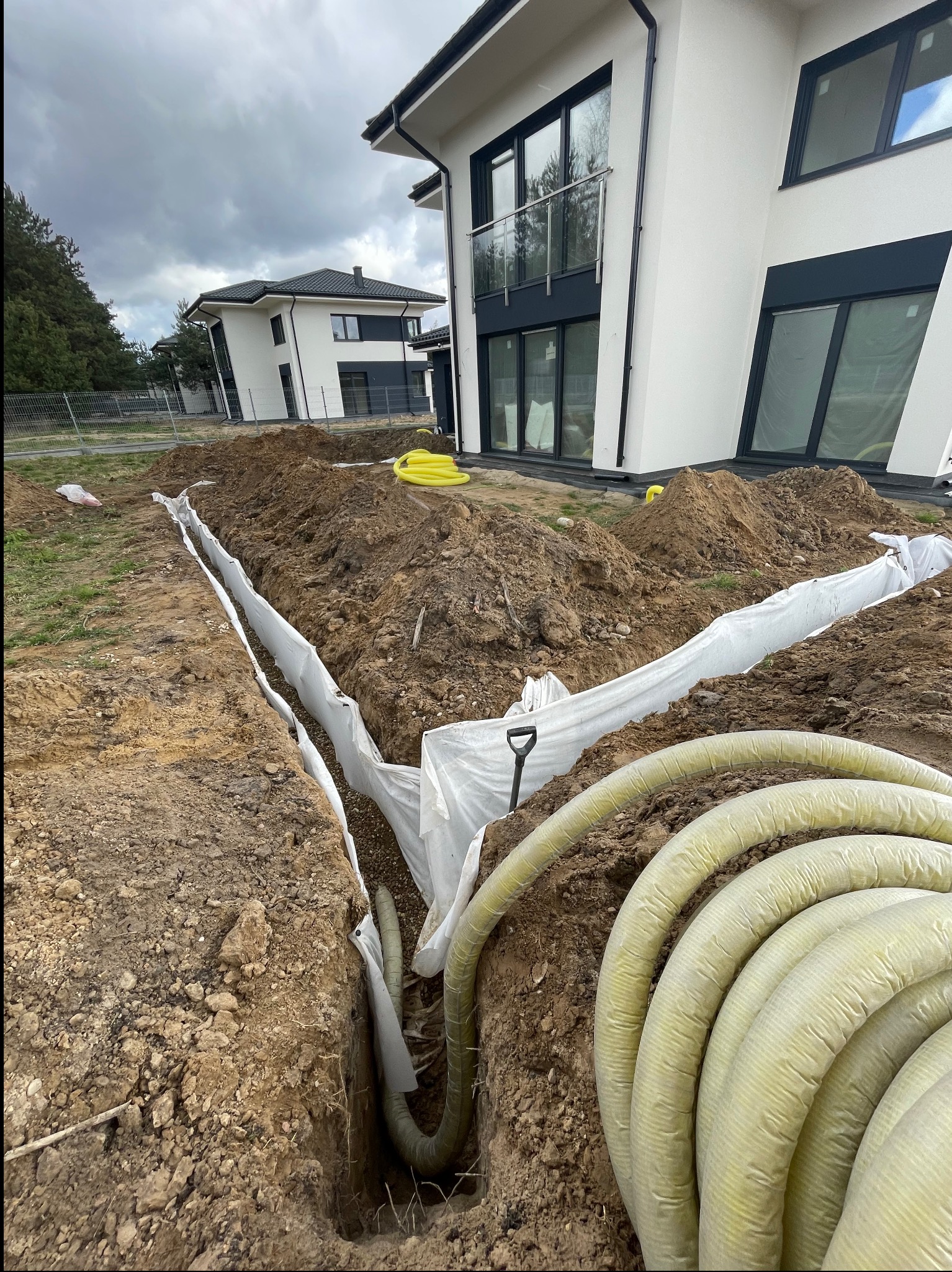 Wykop z rurami drenażowymi w białej geowłókninie na tle nowoczesnego domu w trakcie budowy. Żółte rury w zwojach leżą obok wykopu.