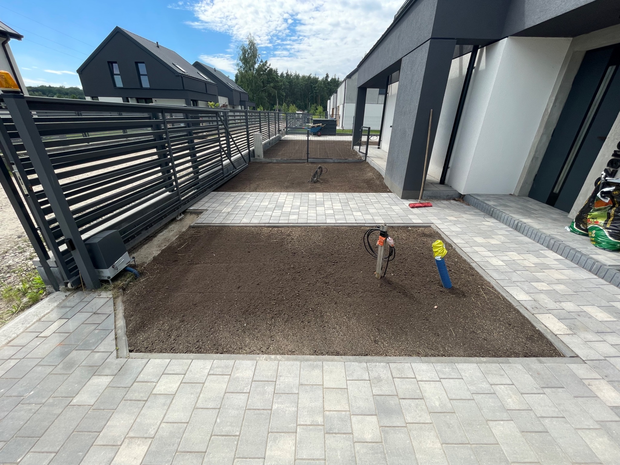 Przygotowany teren pod trawnik z rolki, obrzeża wyłożone kostką brukową. Widoczne wystające rury instalacyjne i świeżo nawieziona ziemia. Nowoczesne budynki w tle.