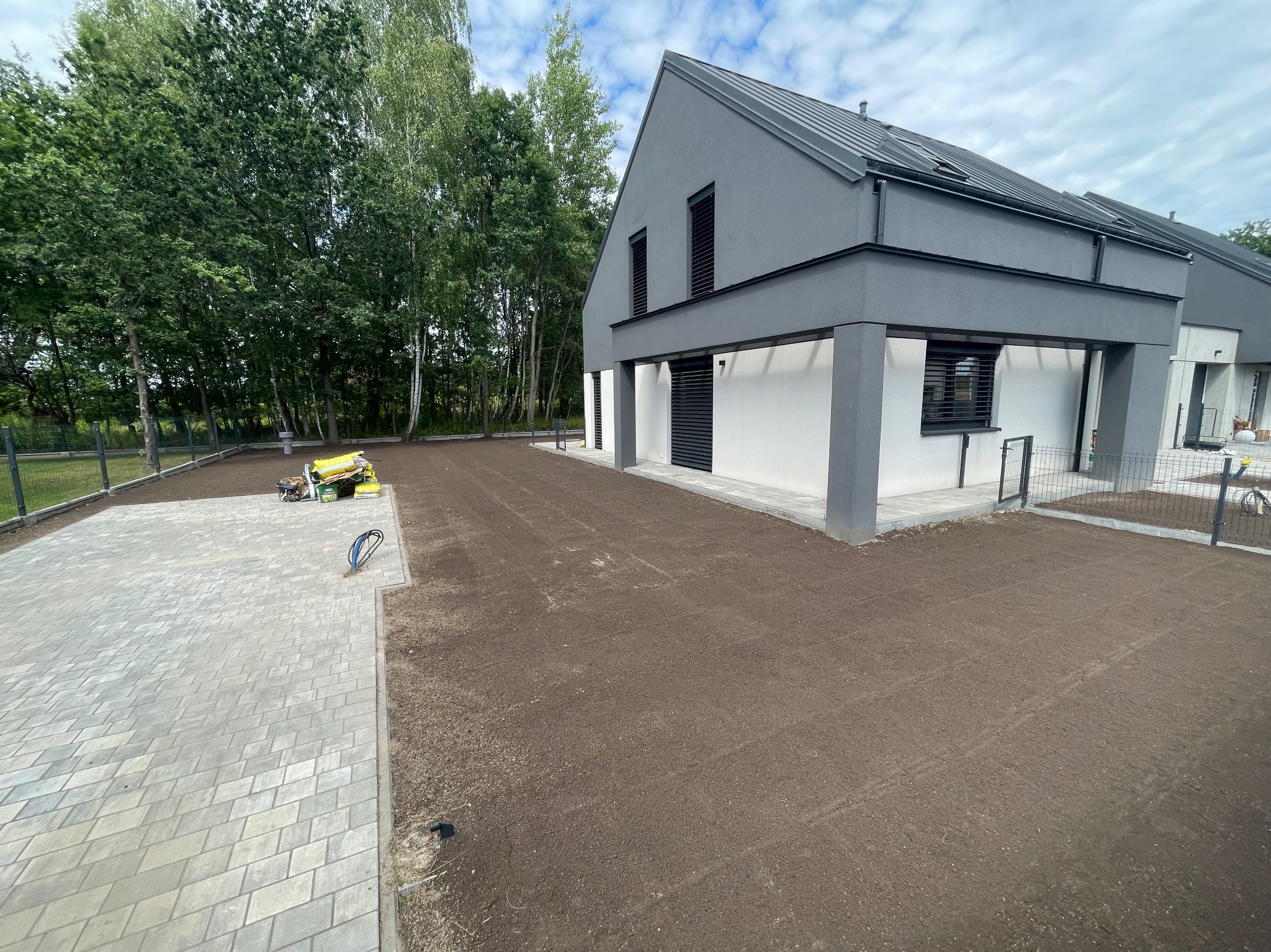 Przygotowany teren pod trawnik z rolki przed nowoczesnym domem. Widoczna ubijarka w tle, wyrównana ziemia i kostka brukowa.