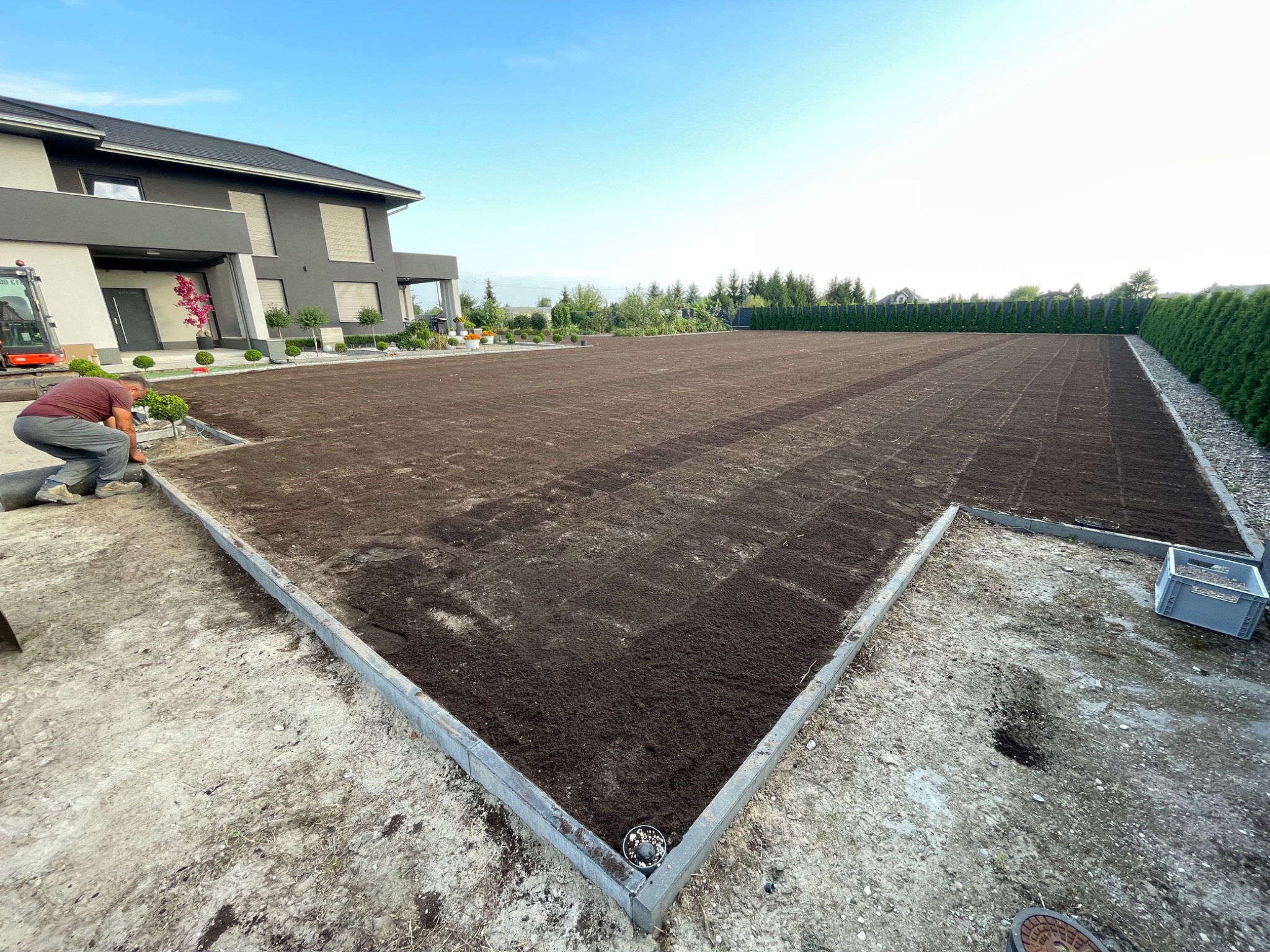 Przygotowany teren pod trawnik z obrzeżami, widoczny pracownik i budynek w tle. Świeżo nawieziona ziemia, gotowa do położenia trawy z rolki. Ujęcie z bliska.