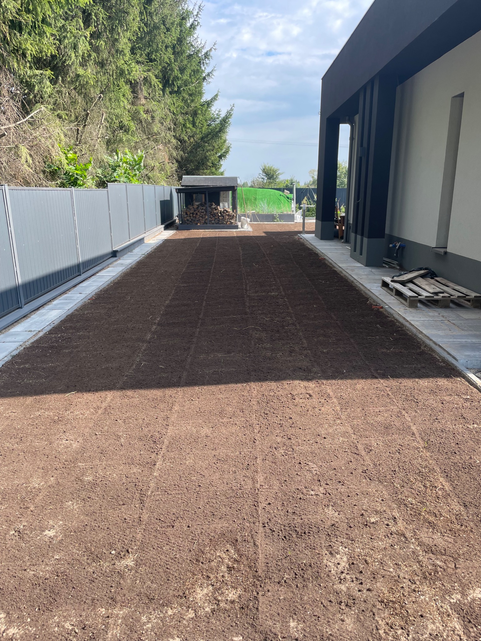 Przygotowany teren pod trawnik z widocznymi śladami po bronowaniu, obrzeża z kostki, szara palisada i budynek z elementami grafitowymi w tle.