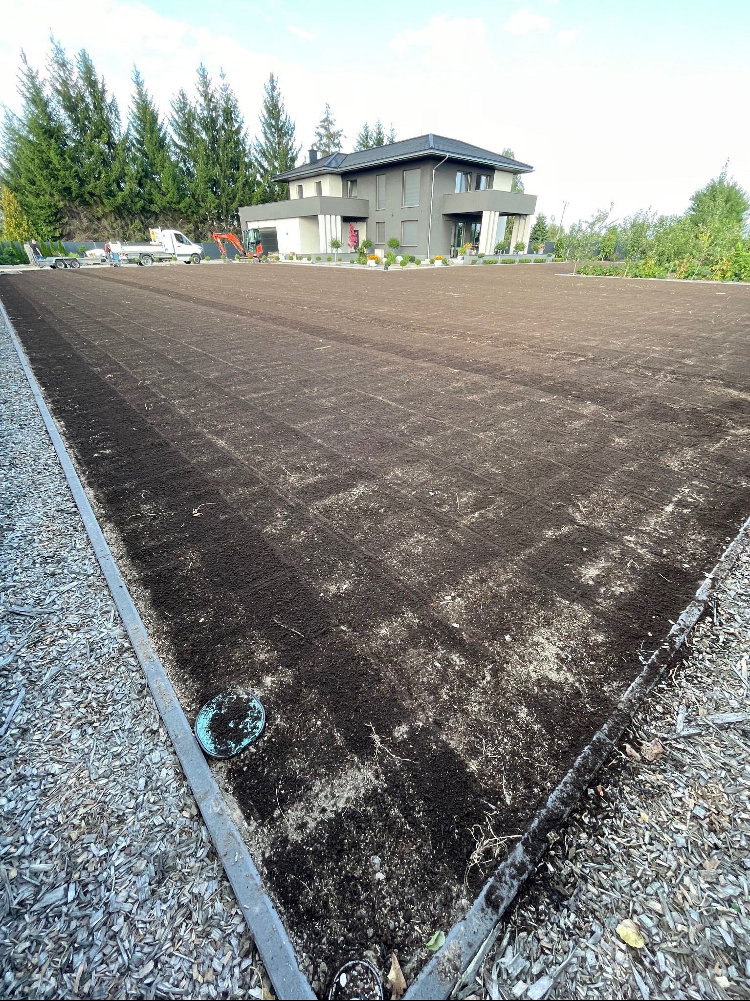 Przygotowany teren pod trawnik z widocznymi śladami po maszynach, nowoczesny dom w tle. Widoczne prace przygotowawcze do założenia trawy.