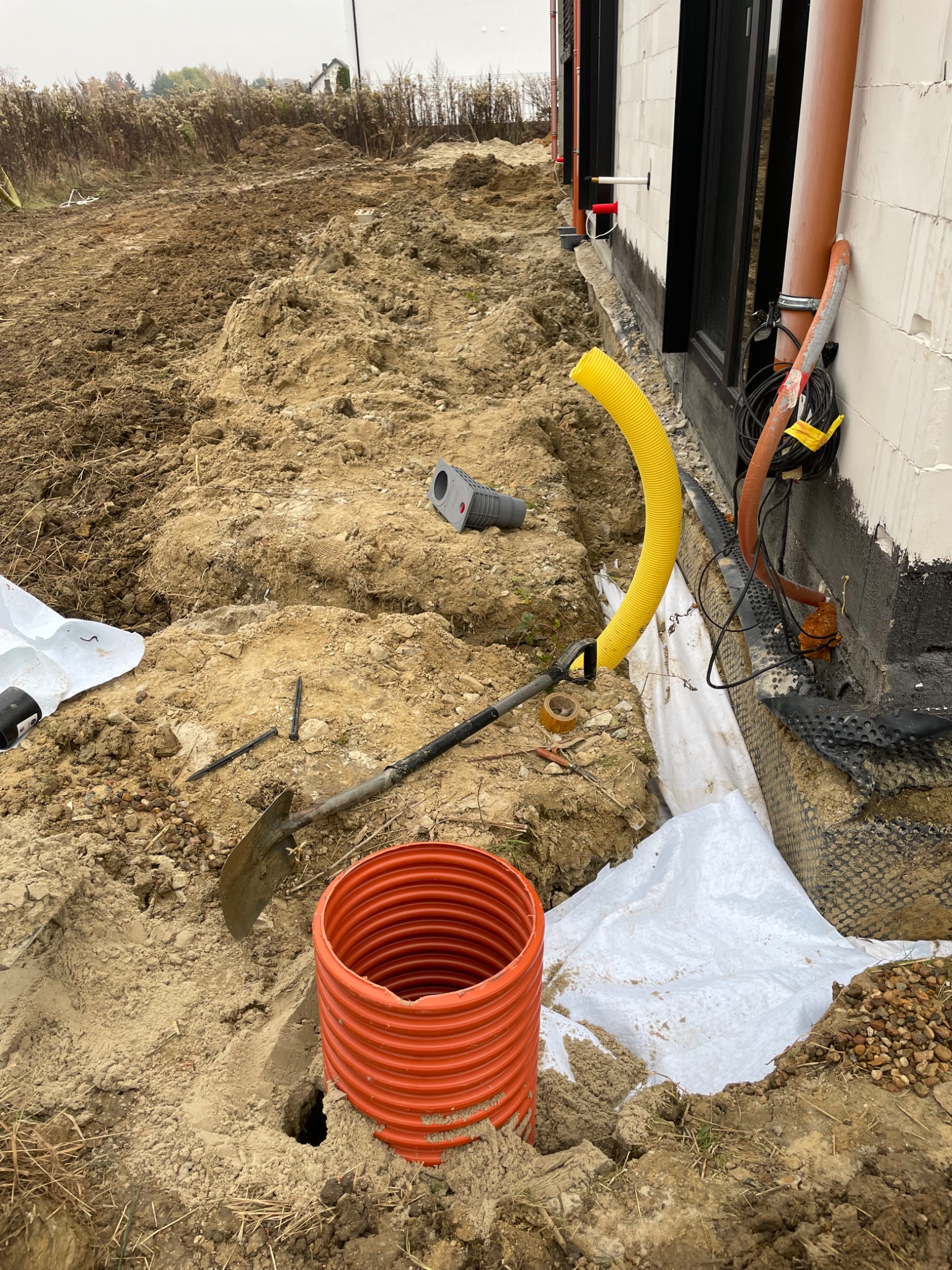 Wykop w ziemi przy fundamencie budynku z widocznymi rurami drenażowymi, folią i narzędziami. Etap prac ziemnych z bliska, w pochmurny dzień.