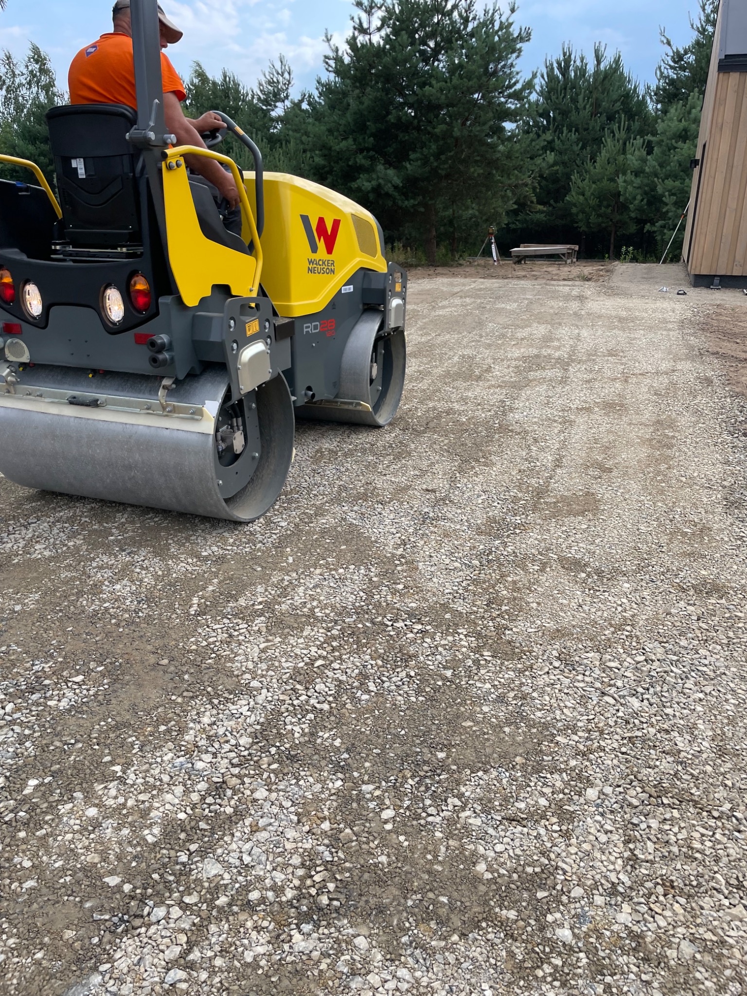 Żółty walec Wacker Neuson RD28 ubija kamienistą nawierzchnię drogi w pobliżu lasu i budynku. Operator w pomarańczowej koszulce siedzi na maszynie.