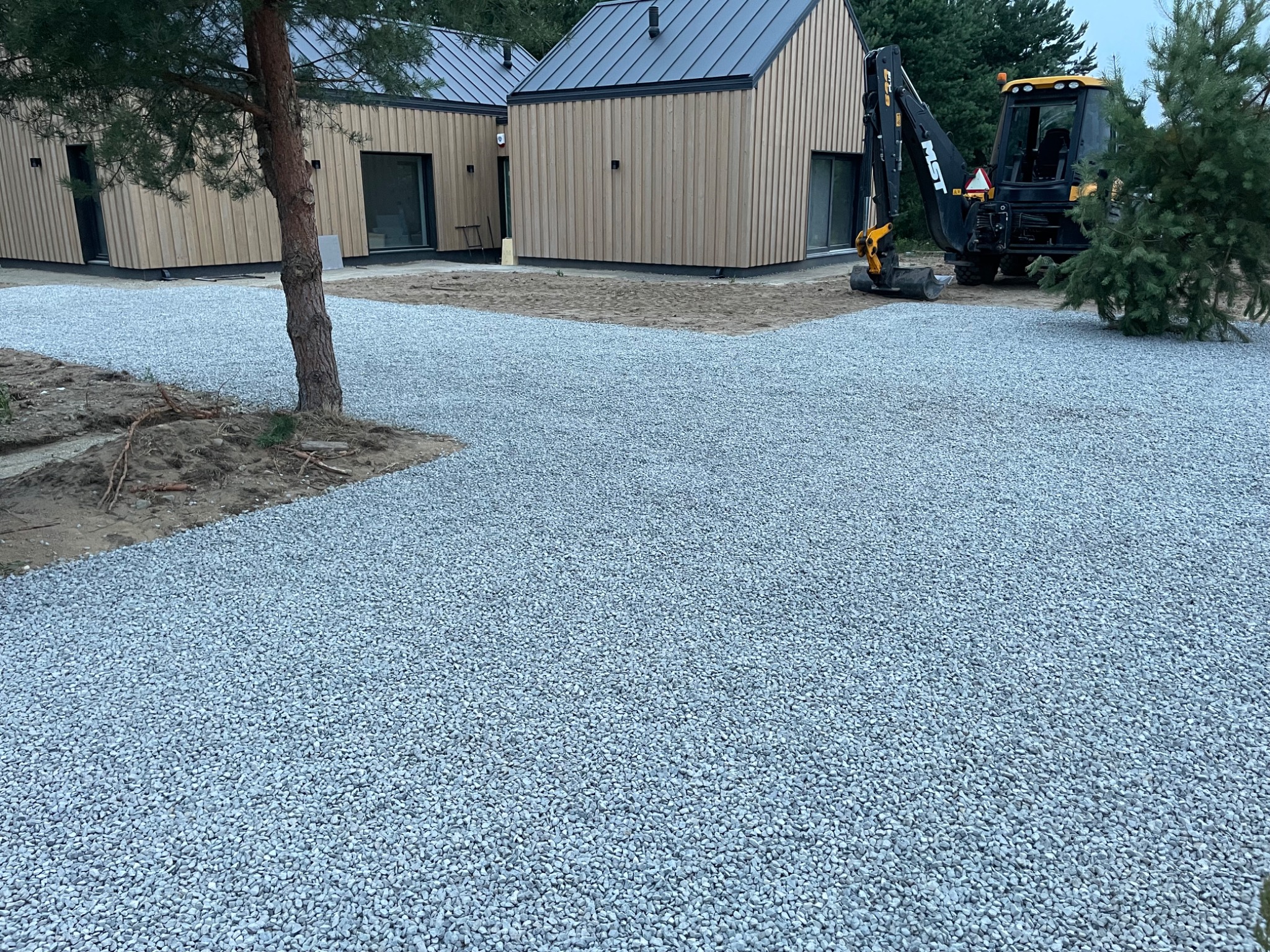 Utwardzona, szara, żwirowa droga do dwóch nowoczesnych domów z drewnianą elewacją. W tle widoczna koparka. Drzewo na pierwszym planie.
