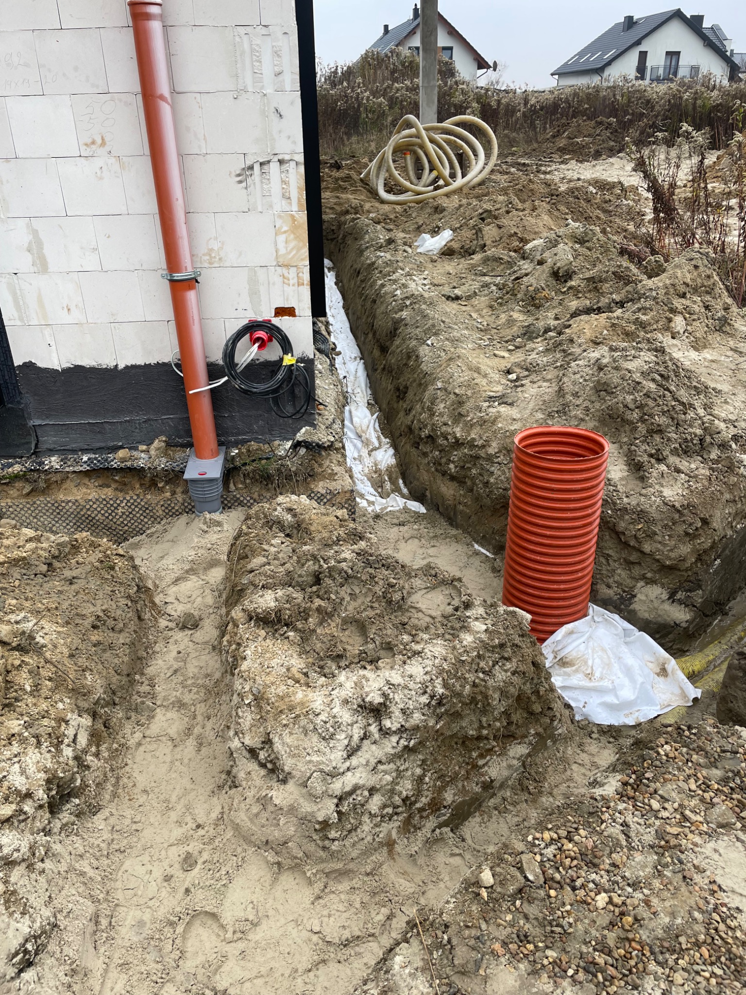 Instalacja rur drenażowych przy fundamencie budynku. Widoczne wykopane rowy, rury odprowadzające wodę i fragment ściany budynku w trakcie budowy.