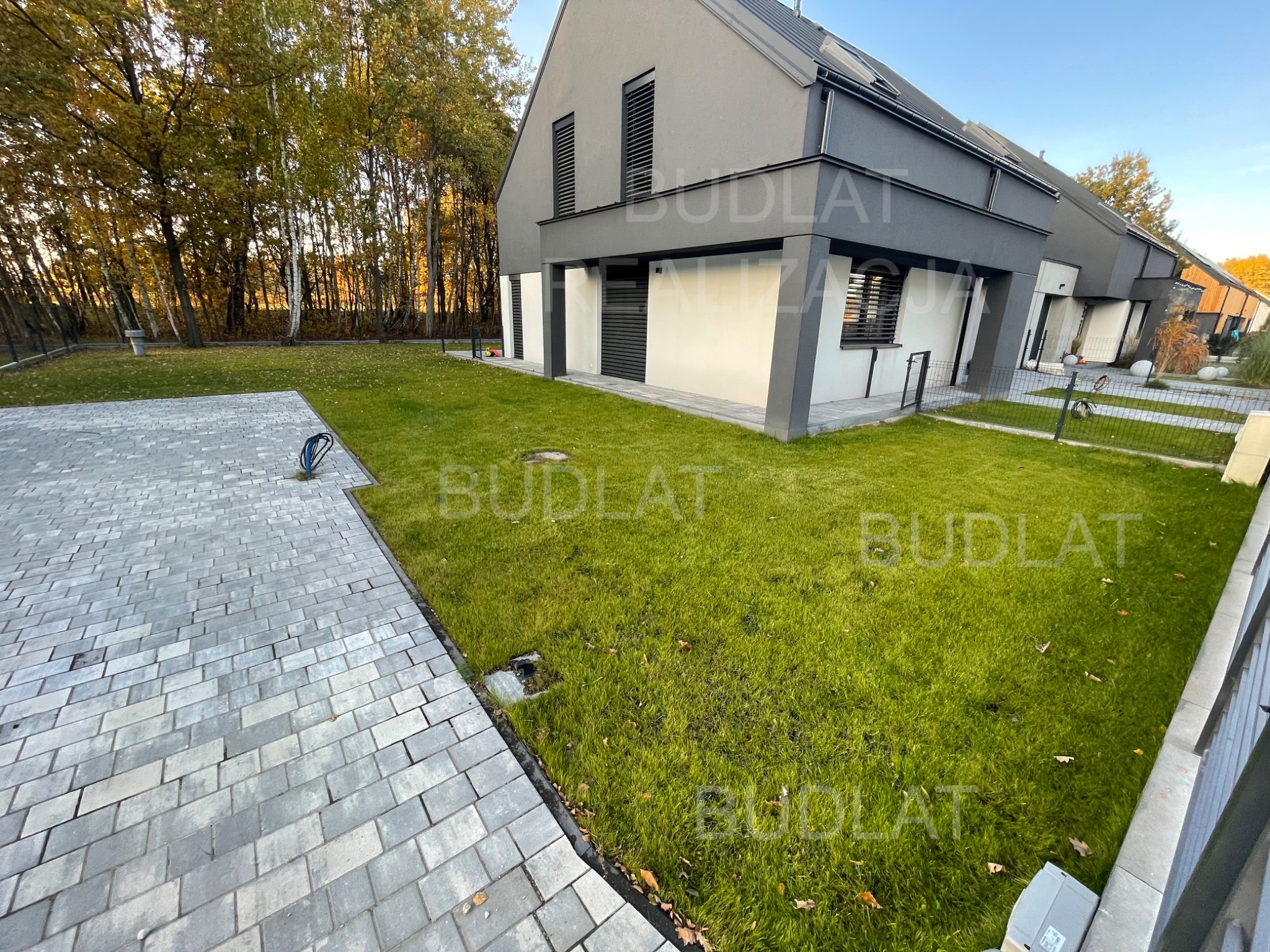 Nowoczesny dom z szarą elewacją i zadbanym trawnikiem. Widoczny fragment brukowanej ścieżki i jesienne drzewa w tle. Ujęcie z poziomu gruntu.