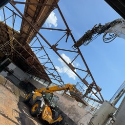 R&C DEMOLUTION SPÓŁKA Z OGRANICZONĄ ODPOWIEDZIALNOŚCIĄ - Żółta ładowarka teleskopowa JCB pracuje wewnątrz hali z odsłoniętą konstrukcją dachu, widok z dołu na błękitne niebo w tle.