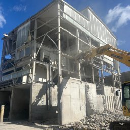 R&C DEMOLUTION SPÓŁKA Z OGRANICZONĄ ODPOWIEDZIALNOŚCIĄ - Rozbiórka budynku z użyciem koparki Komatsu. Fragment konstrukcji w trakcie demontażu, widoczne gruzowisko i niebo z chmurami. Dzień, jasne oświetlenie.