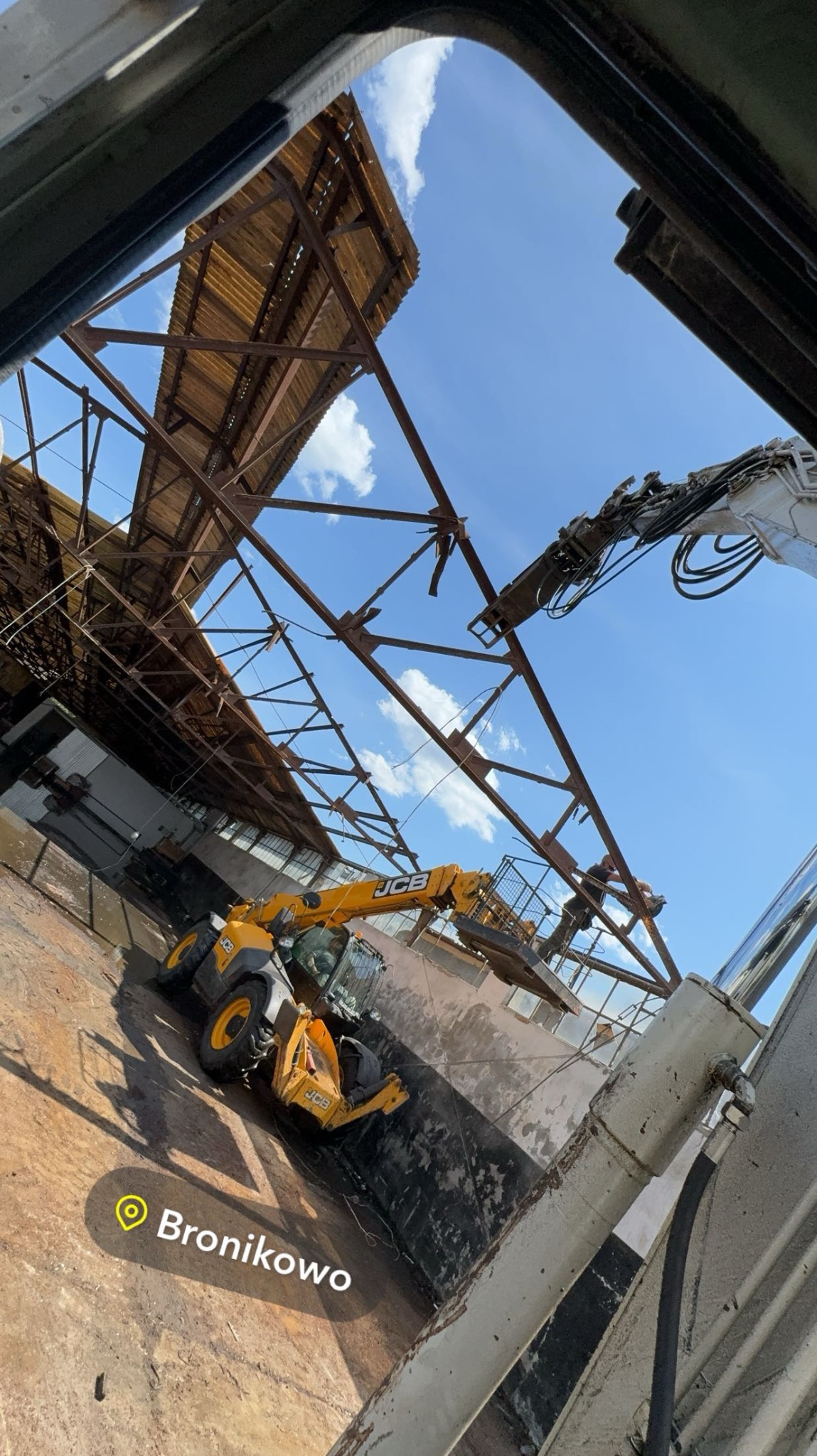 Żółta ładowarka teleskopowa JCB pracuje wewnątrz hali z odsłoniętą konstrukcją dachu, widok z dołu na błękitne niebo w tle.