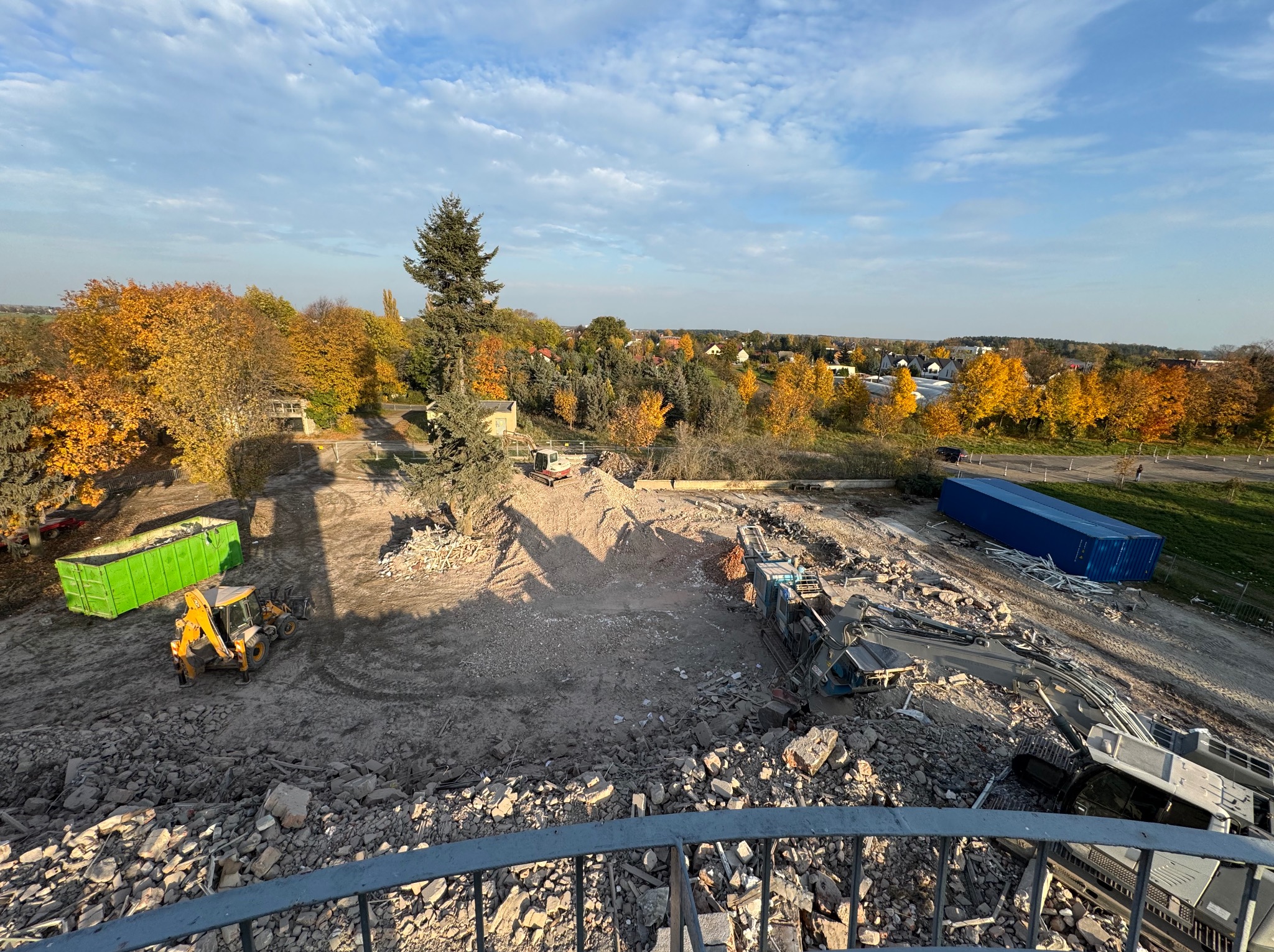 Widok z góry na plac budowy w Poznaniu po rozbiórce, z gruzem, kontenerami i maszynami budowlanymi na tle jesiennego krajobrazu.