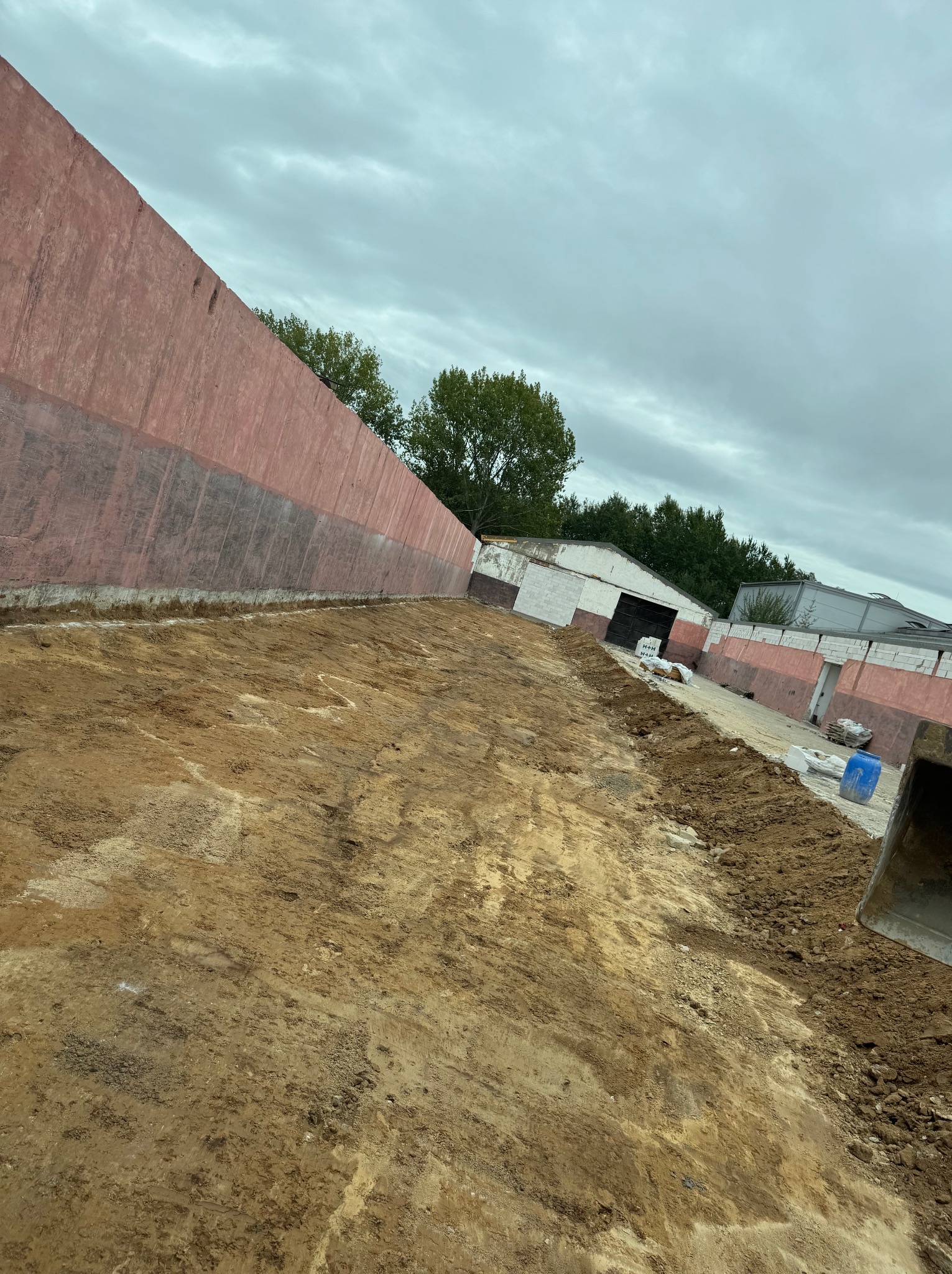 Przygotowany teren pod budowę, widoczna wykopana ziemia, fragment ściany budynku w tle i fragment łyżki koparki po prawej stronie kadru.