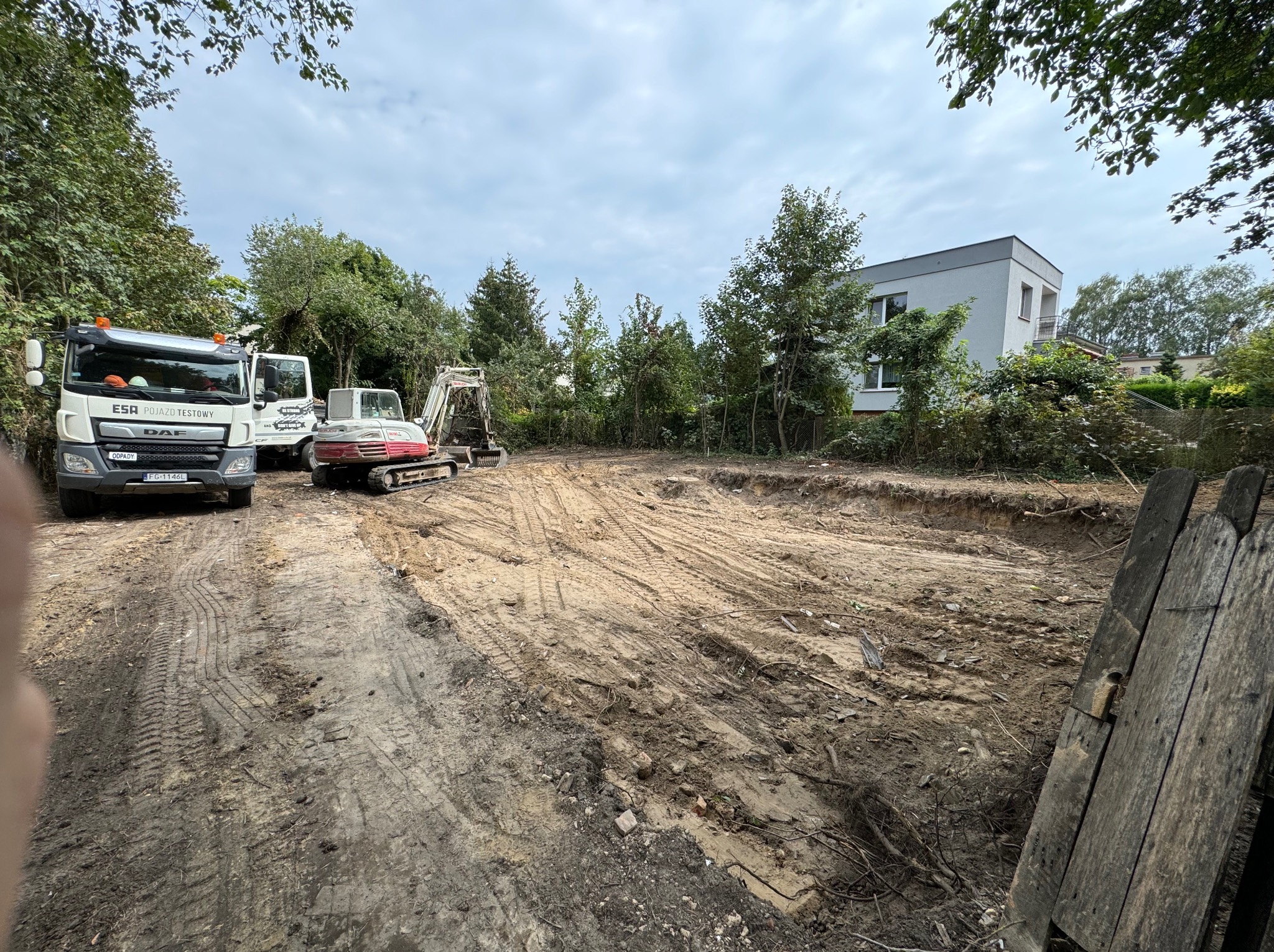 Przygotowany teren pod budowę z widocznymi śladami opon, koparką, dwoma samochodami ciężarowymi oraz fragmentem drewnianego ogrodzenia w prawym dolnym rogu.