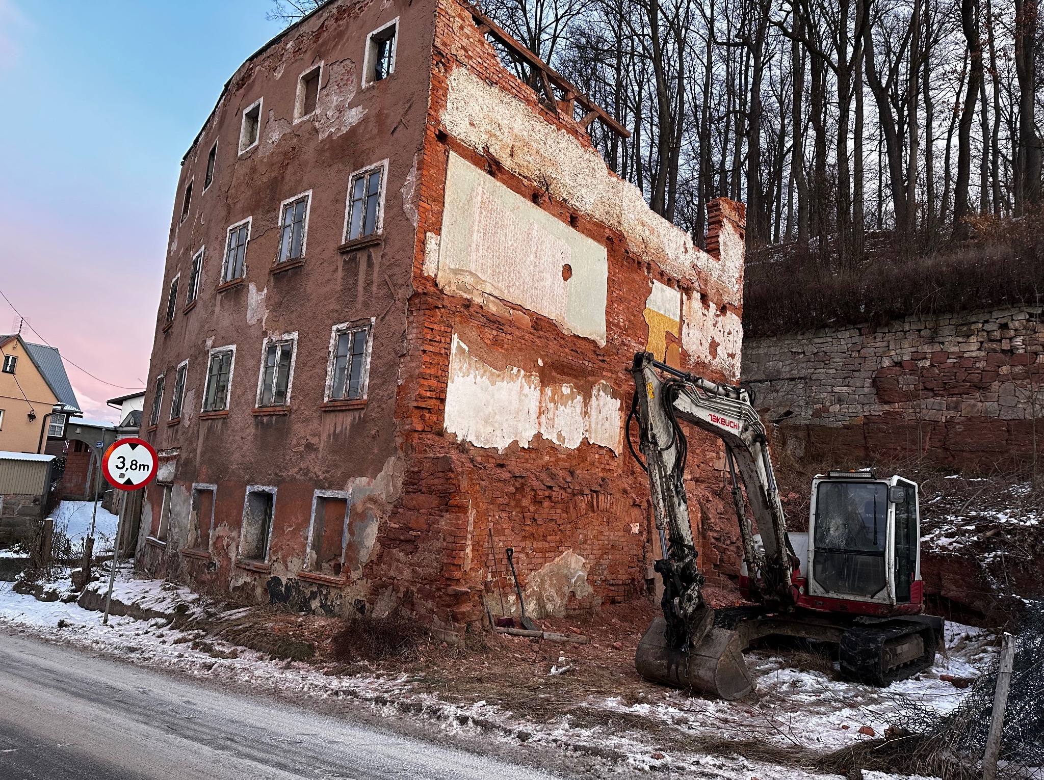 Częściowo rozebrany, ceglany budynek z pustymi otworami okiennymi, obok którego stoi mała koparka Takeuchi, widoczny znak drogowy ograniczający wysokość na tle zimowego krajobrazu.