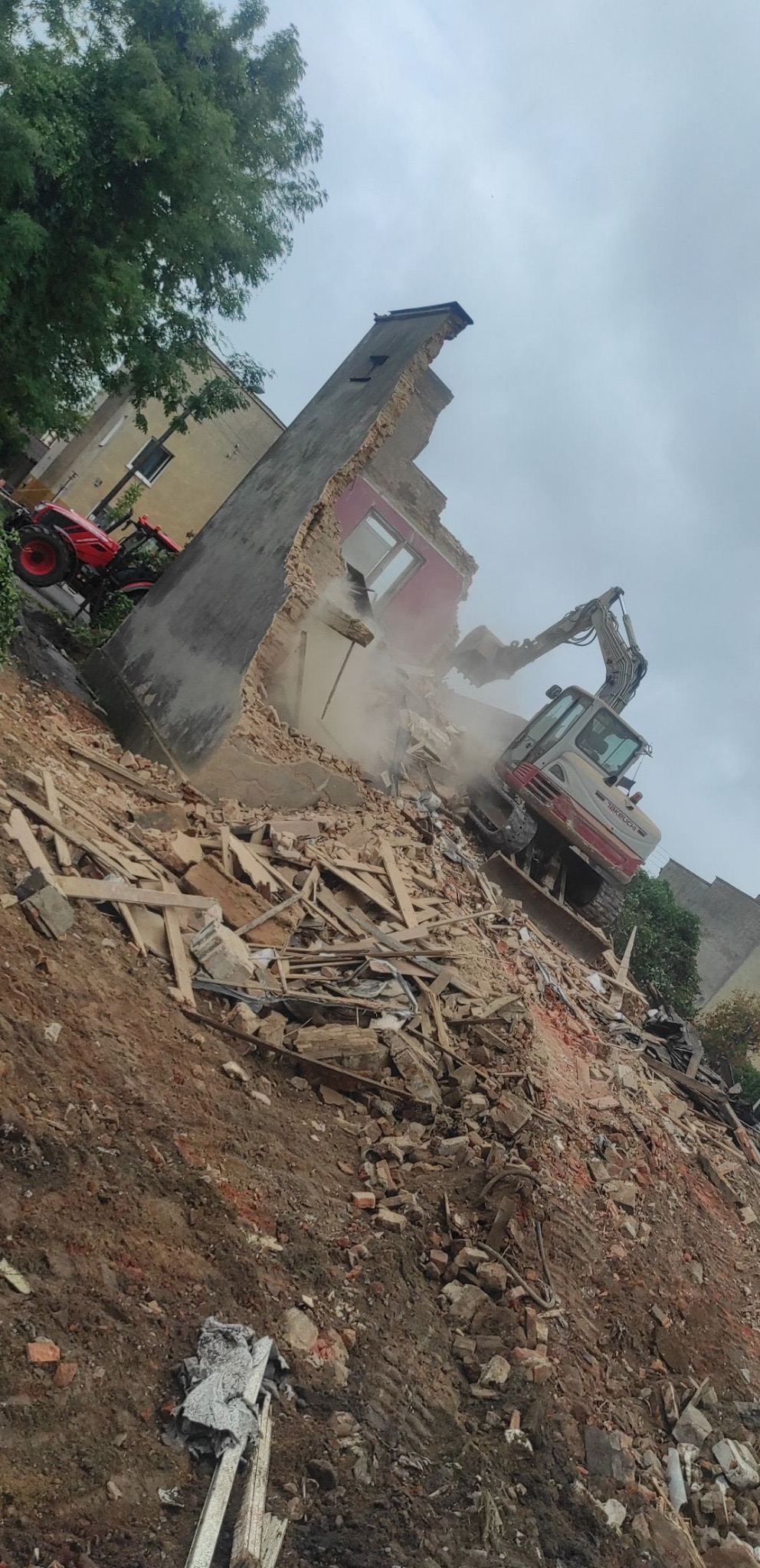 Częściowo zburzony budynek z odsłoniętym czerwonym pokojem, gruzowisko i pracująca koparka Takeuchi na zboczu, czerwony traktor w tle.