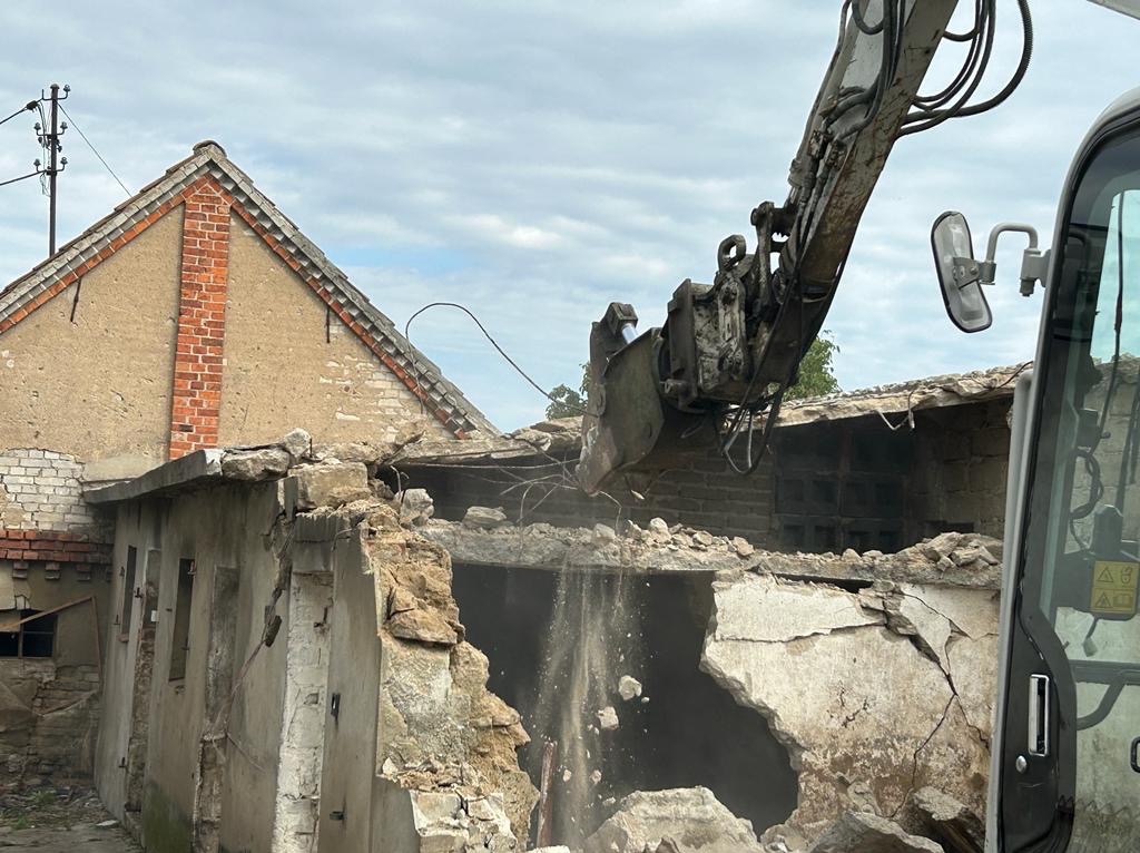 Koparka z łyżką w trakcie kruszenia muru starego, częściowo zburzonego budynku z ceglanym wykończeniem dachu, widok z kabiny operatora.