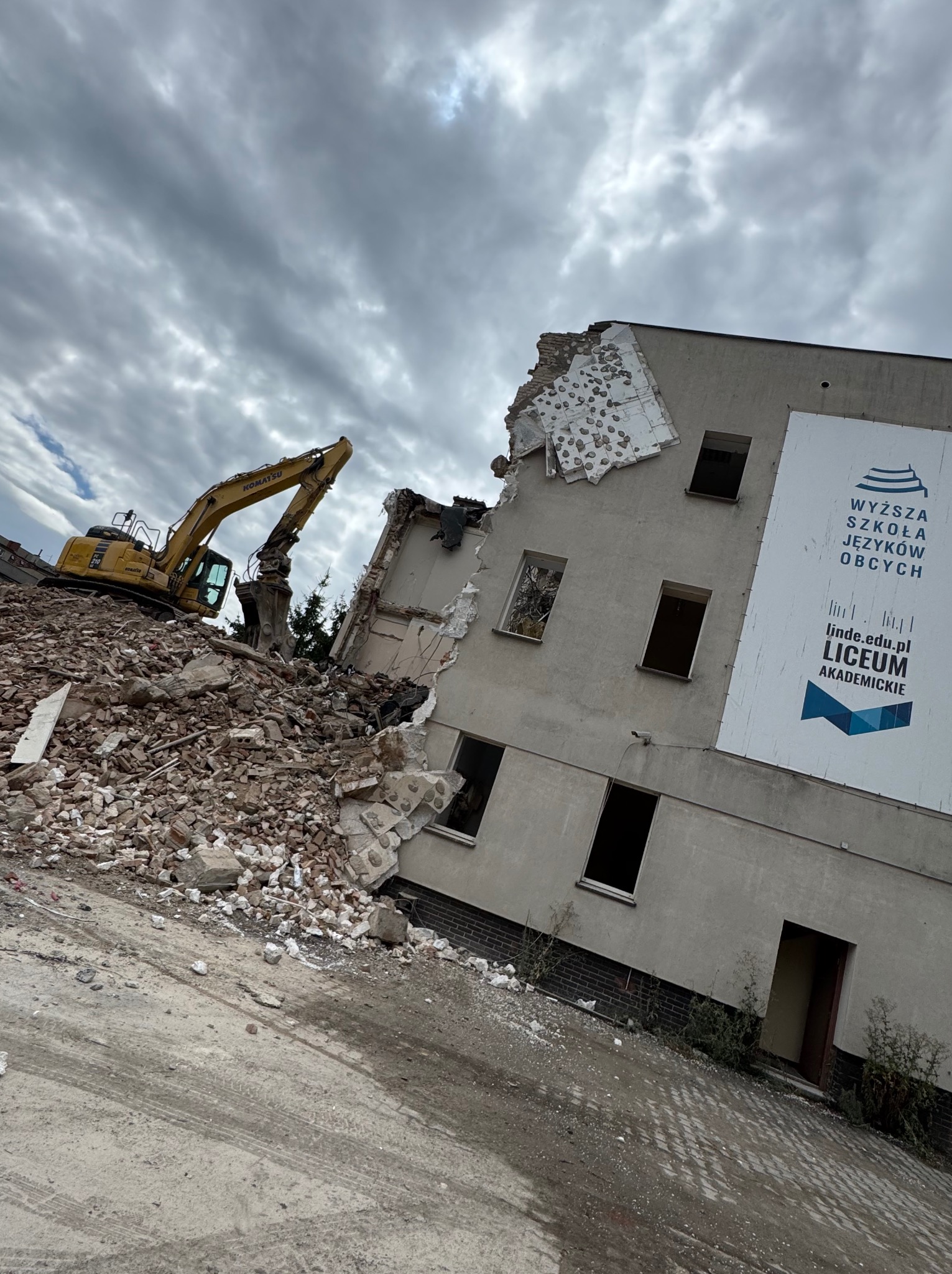 Żółta koparka Komatsu na gruzowisku, częściowo zburzony budynek z szyldem 'Wyższa Szkoła Języków Obcych Liceum Akademickie', pochmurne niebo.