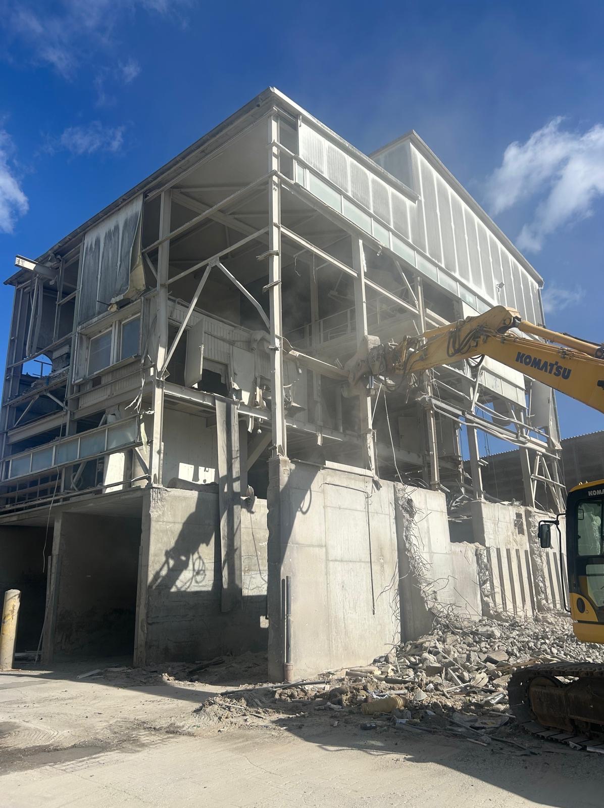 Rozbiórka budynku z użyciem koparki Komatsu. Fragment konstrukcji w trakcie demontażu, widoczne gruzowisko i niebo z chmurami. Dzień, jasne oświetlenie.
