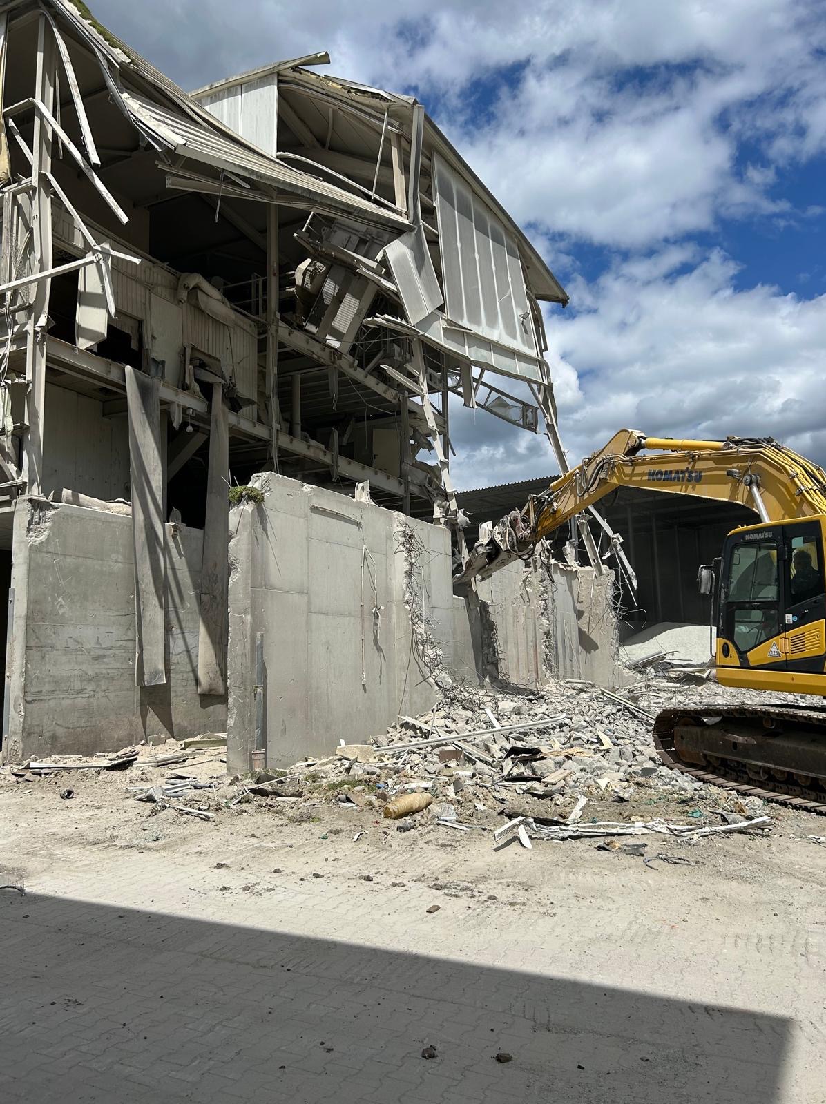 Żółta koparka Komatsu podczas rozbiórki budynku przemysłowego. Widoczne zniszczenia i gruz. Niebieskie niebo z chmurami w tle. Wysoki kadr.