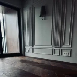 Ułożona w jodełkę, ciemna, drewniana podłoga w nowocześnie urządzonym wnętrzu z szarą ścianą, dekoracyjnymi listwami i czarnym oknem balkonowym.