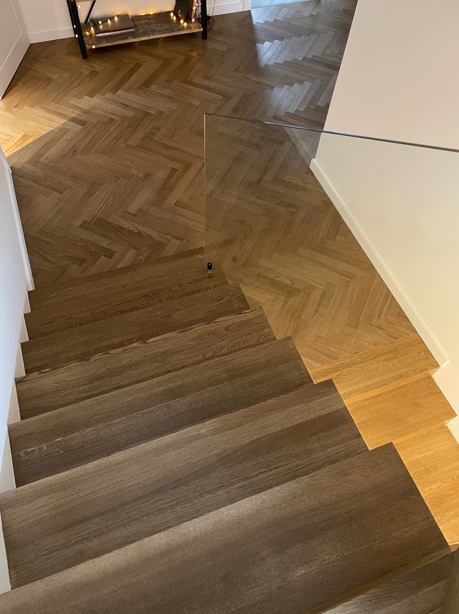 Ukończone schody z parkietu ułożonego w jodełkę, łączące się z podłogą w tym samym wzorze, zabezpieczone szklaną balustradą w domu w Warszawie.