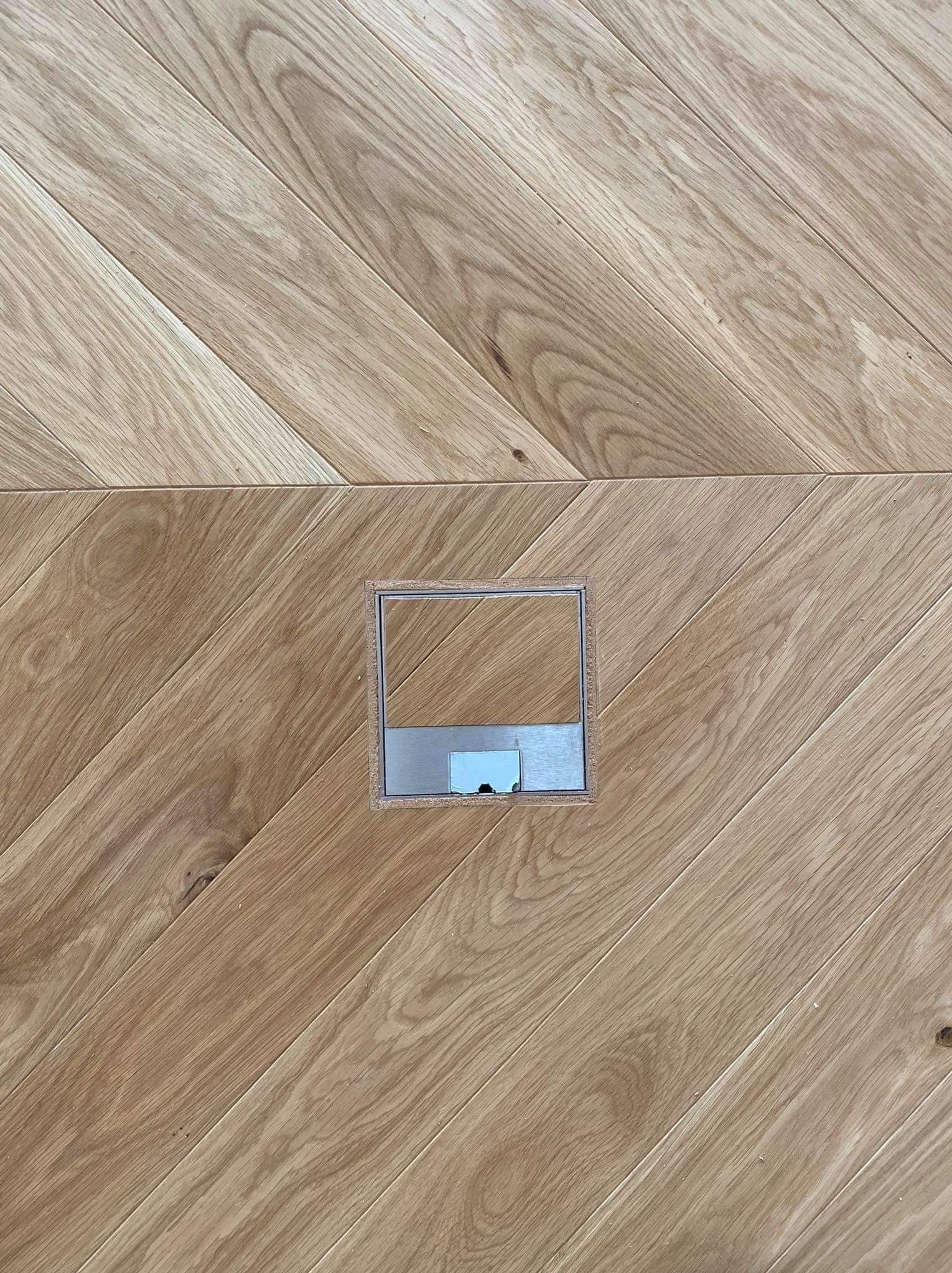 Ułożona w jodełkę jasna podłoga z drewna z metalową puszką podłogową na gniazdka.