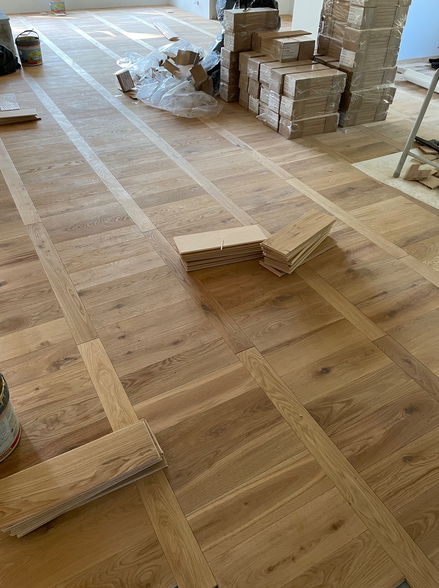 Układanie parkietu w jodełkę francuską, widoczne ułożone fragmenty i paczki paneli w tle, przygotowane do montażu.