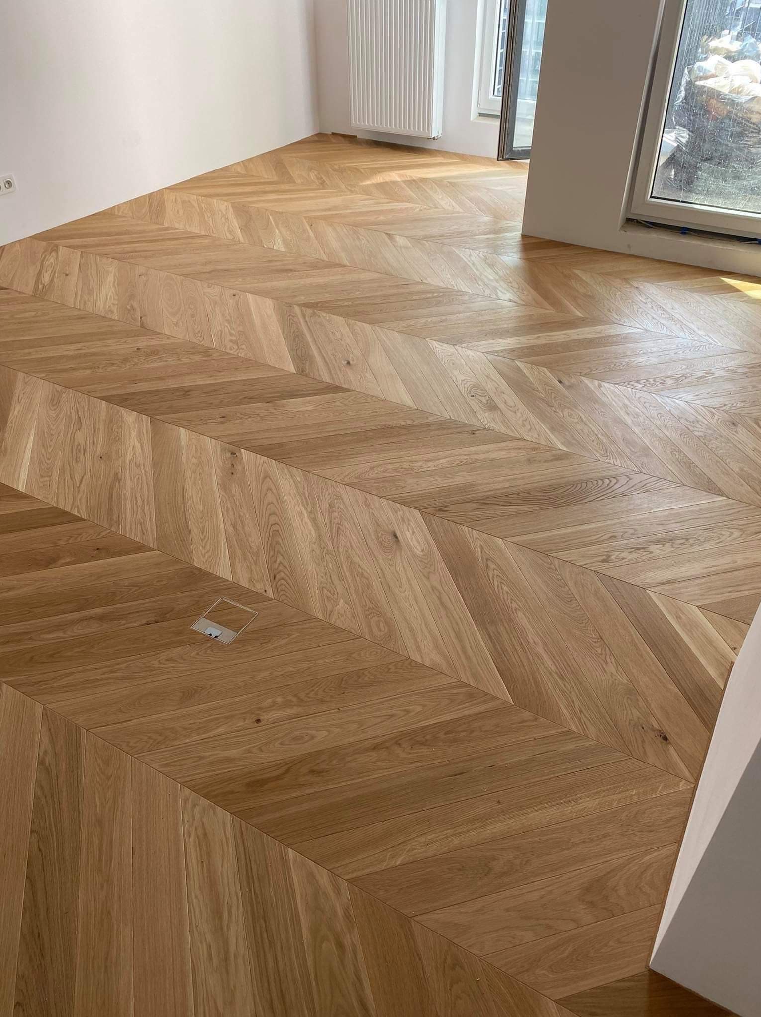 Parkiet ułożony we wzór jodełki francuskiej, jasne drewno dębowe, widoczny fragment gniazdka podłogowego, jasne ściany i okno w tle.