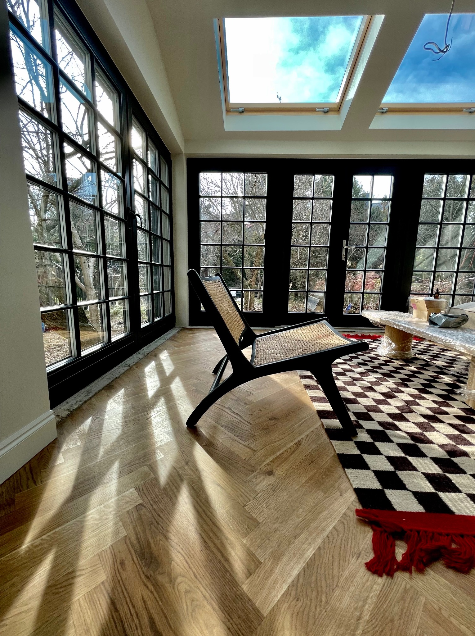 Jasna podłoga drewniana ułożona we wzór jodełki francuskiej w nowocześnie urządzonym salonie z dużymi oknami i świetlikami dachowymi, na podłodze leży czarno-biały dywan w szachownicę, a obok stoi...