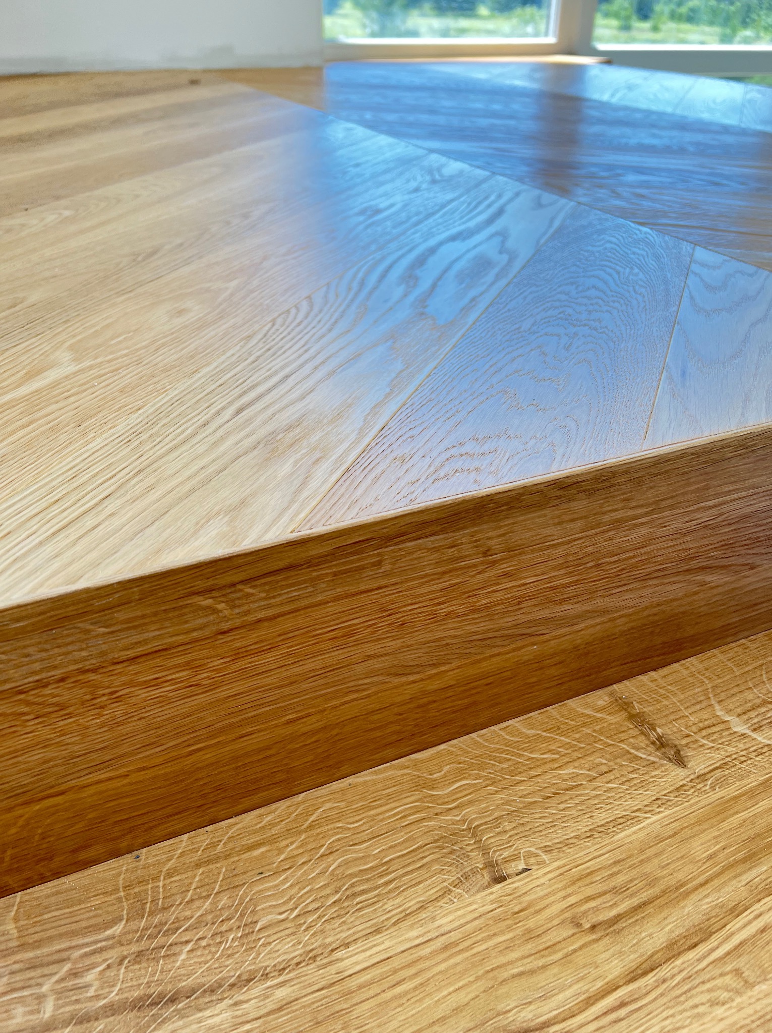 Jasna, lakierowana podłoga drewniana z wyraźnym usłojeniem, widoczny fragment progu i okno w tle.